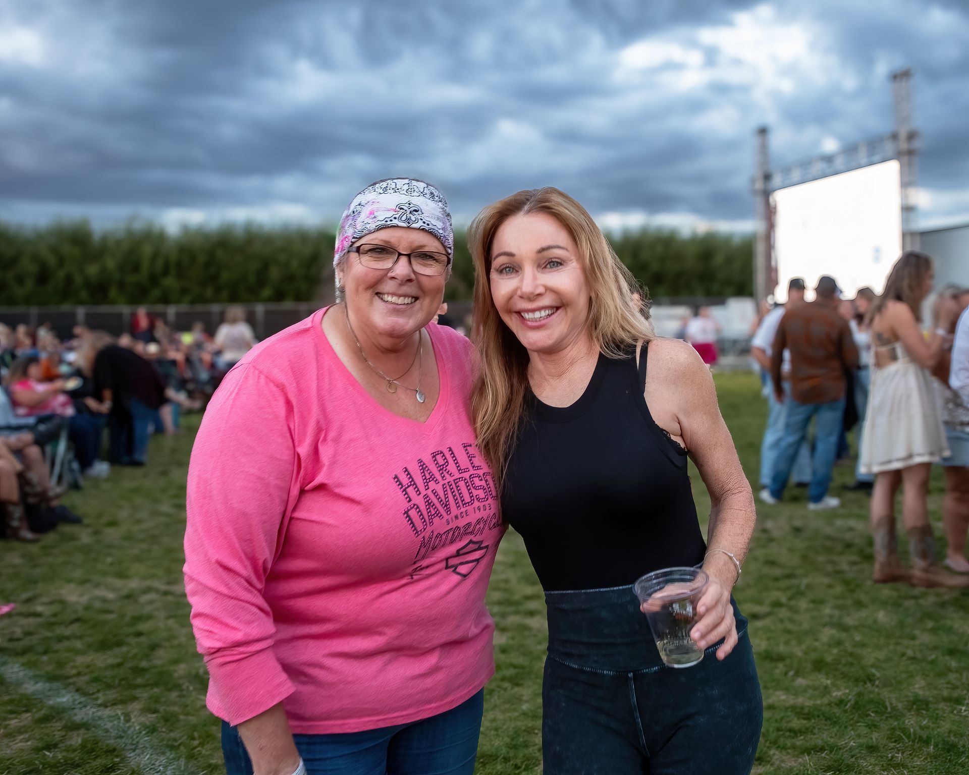 Two women smiling, one in pink Harley Davidson shirt, the other in black, at an outdoor event.