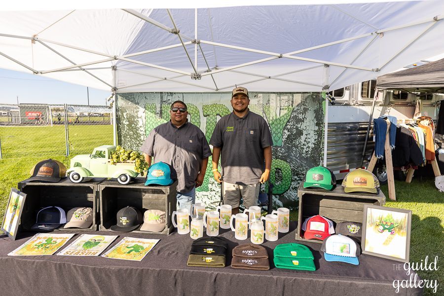 Two men are standing in front of a table filled with hats and mugs.