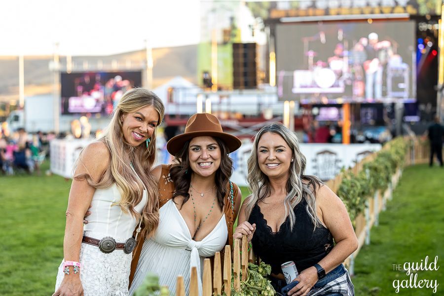 Three women are posing for a picture next to a wooden fence at a concert.