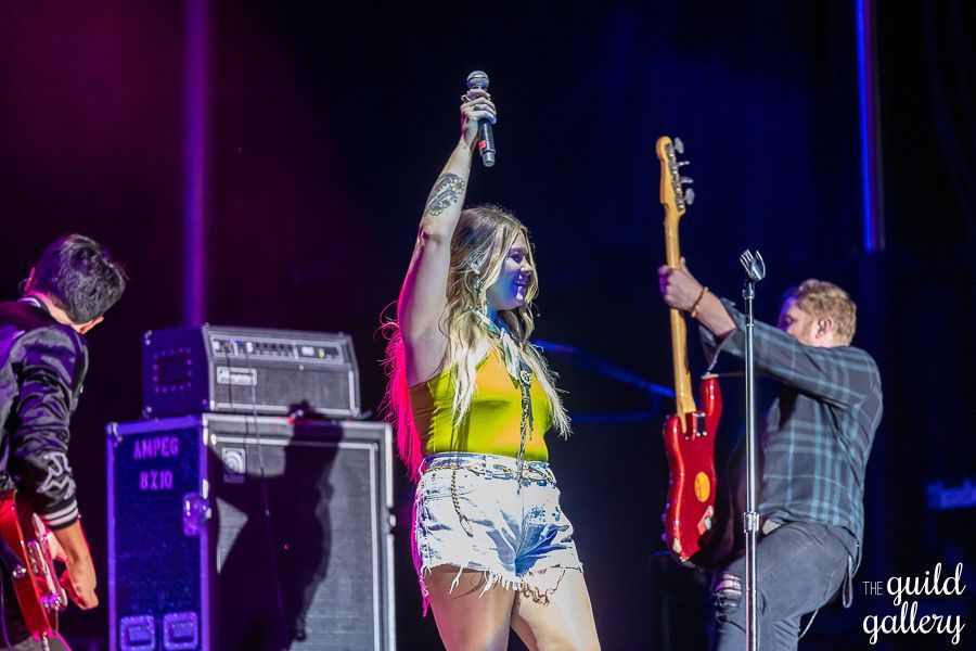 A woman is singing into a microphone on a stage while two men play guitars.