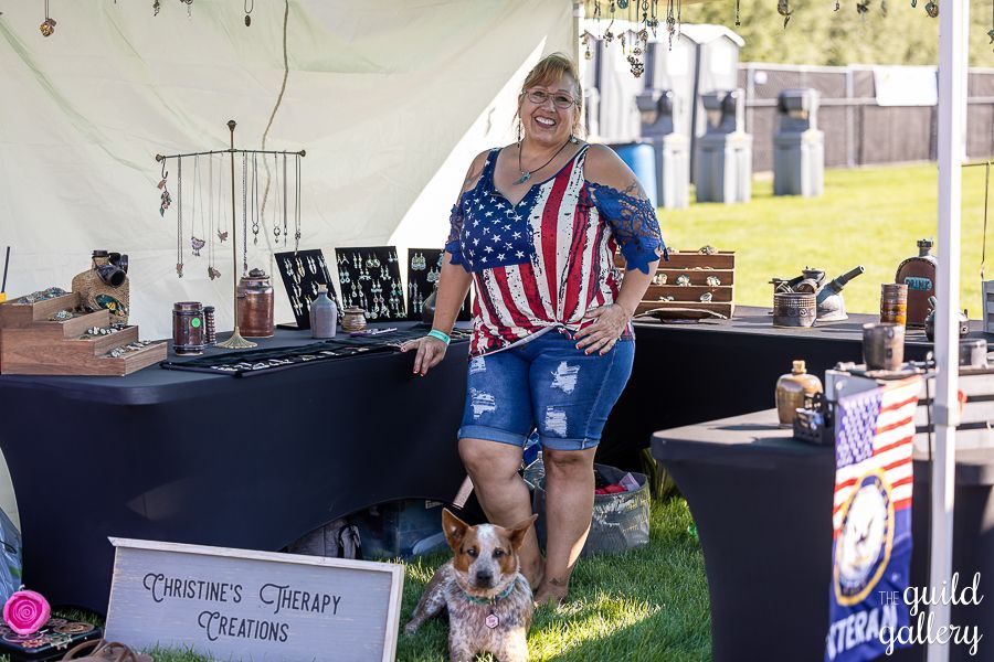 A woman in an american flag shirt is standing next to a dog in front of a table.