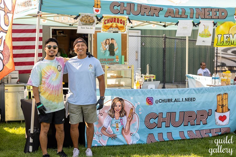 Two men are standing in front of a churro stand at a festival.