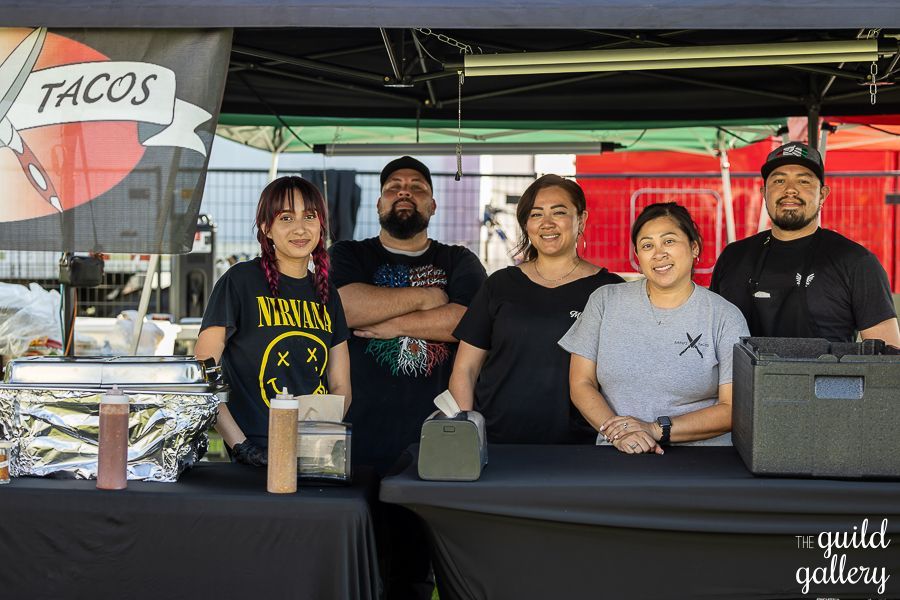 A group of people standing in front of a tacos stand.
