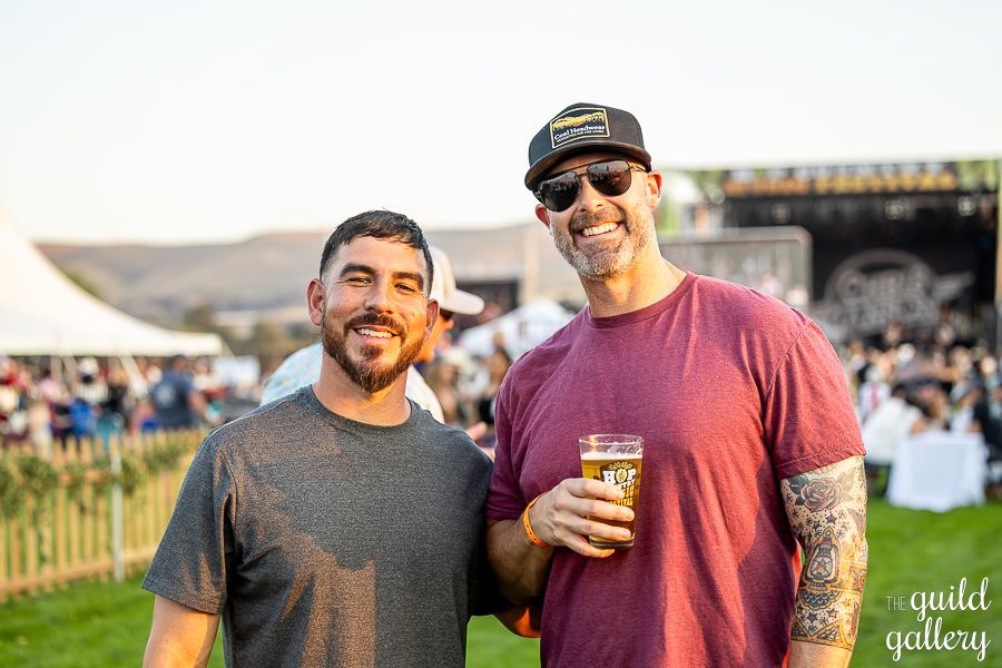 Two men are standing next to each other in a field holding glasses of beer.