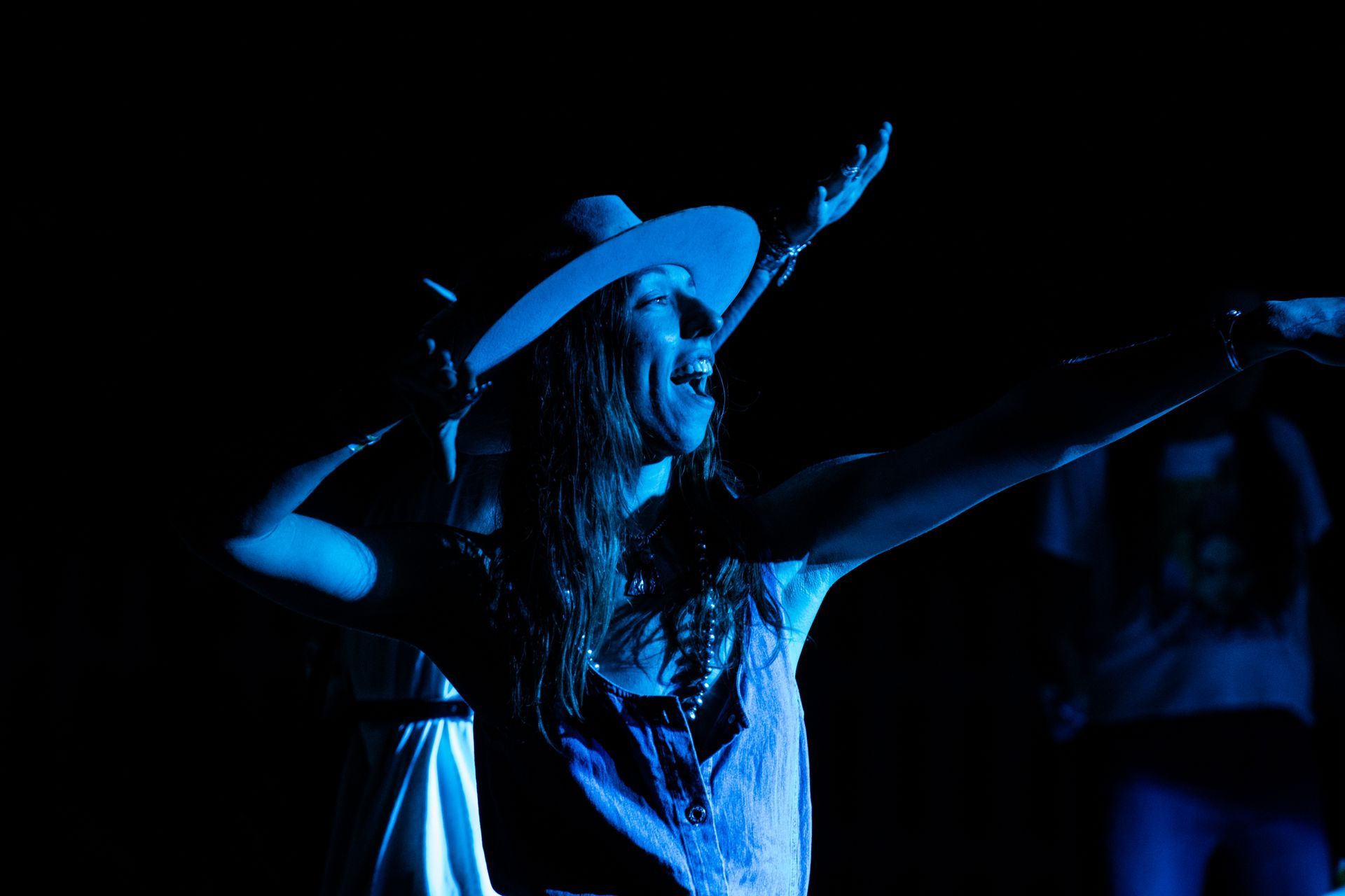 A woman in a cowboy hat is standing in the dark with her arms outstretched.