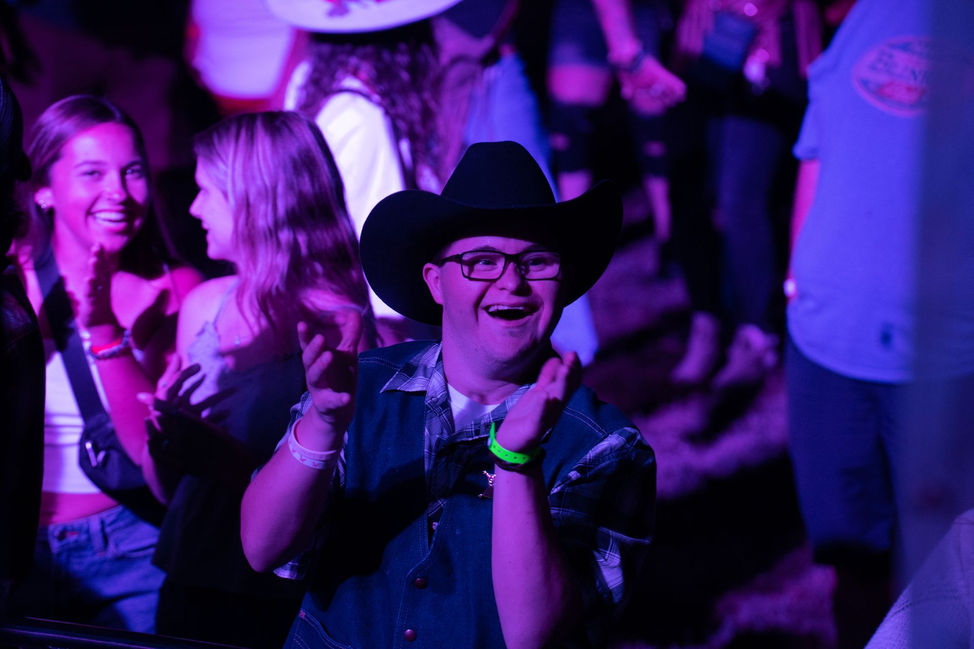 A man wearing a cowboy hat and glasses is sitting in a crowd of people at a concert.