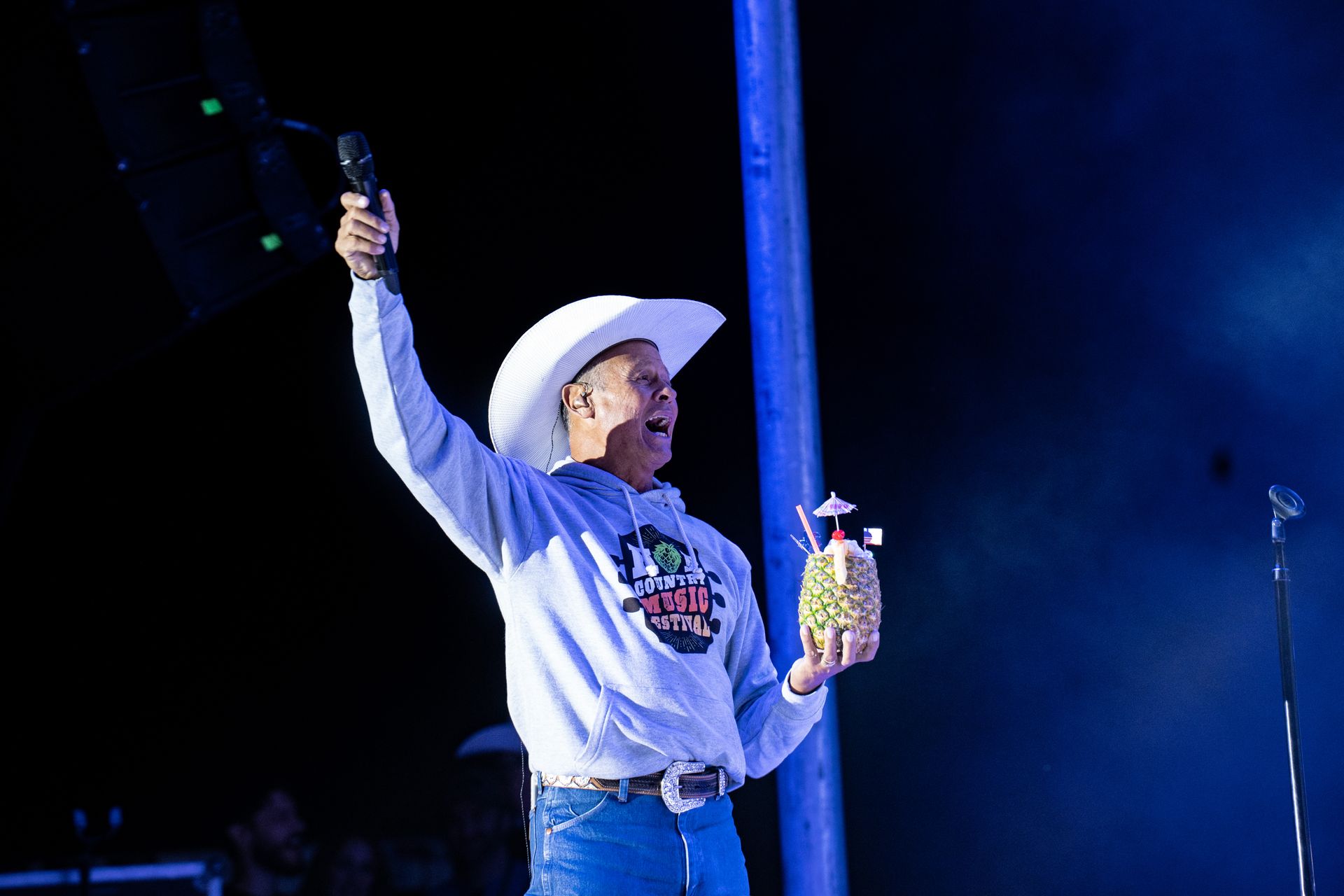 A man in a cowboy hat is holding a microphone and a pineapple.