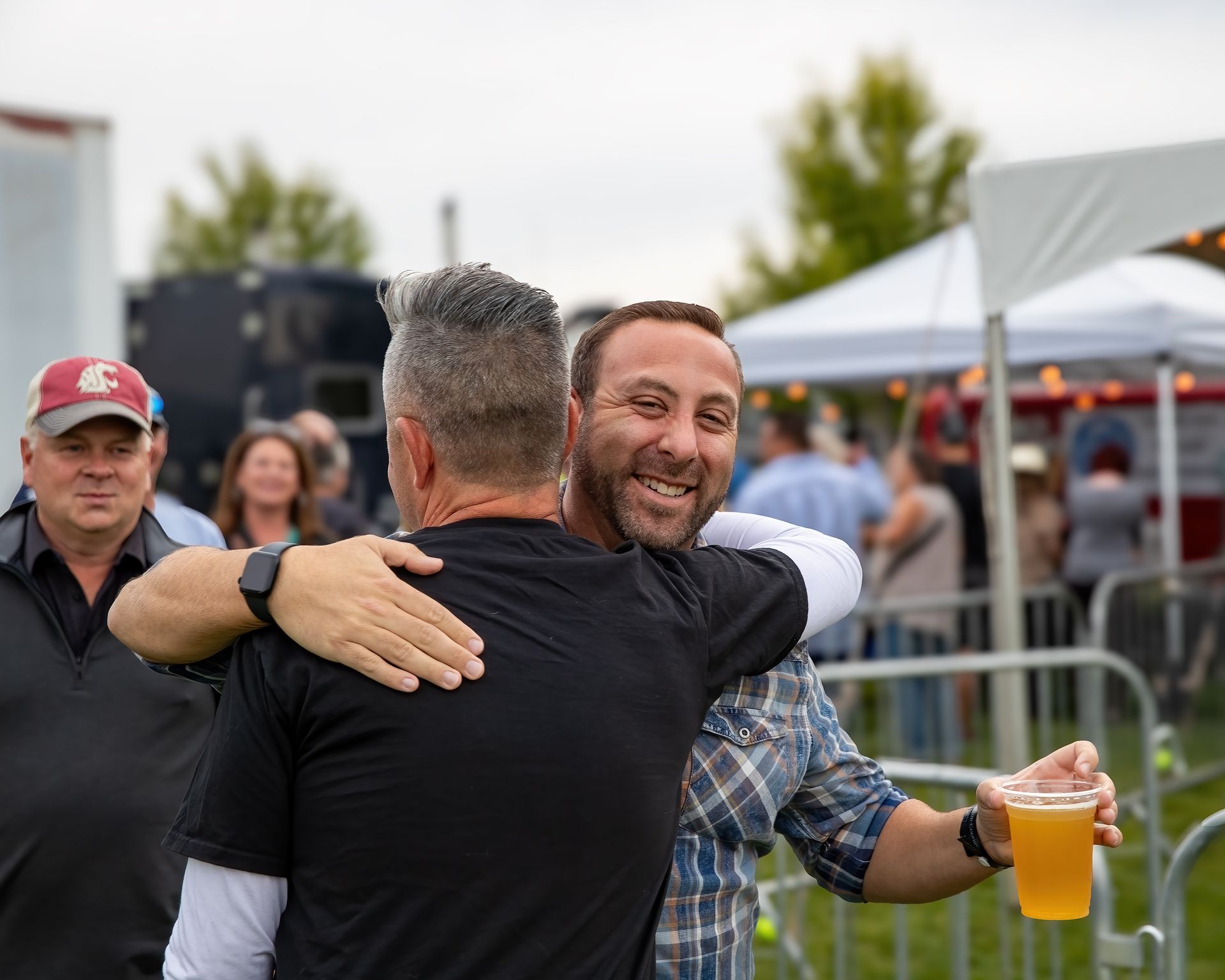 Two men are hugging each other at a festival and one of them is holding a glass of beer.