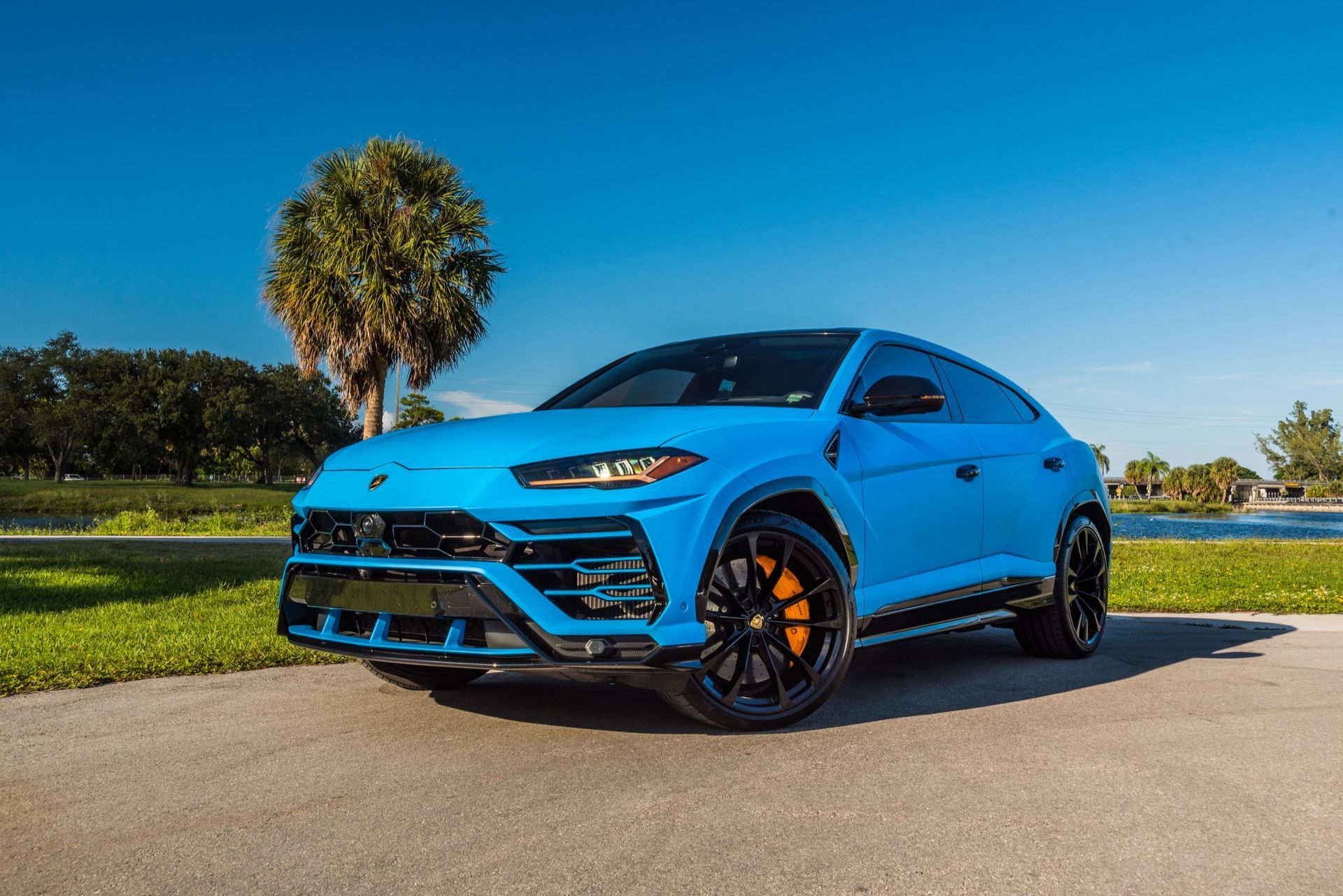 Blue Lamborghini Urus SUV parked on a paved road with a lake and trees in the background.
