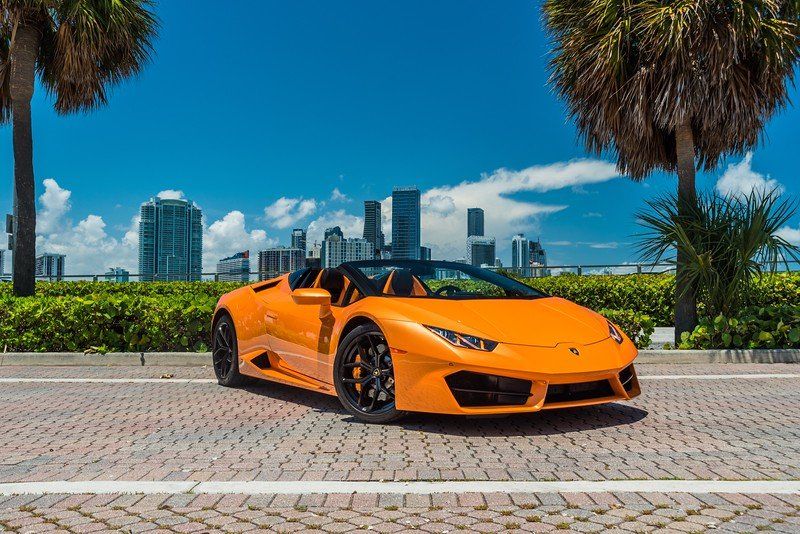 Orange Lamborghini convertible parked on brick road with city skyline and palm trees.