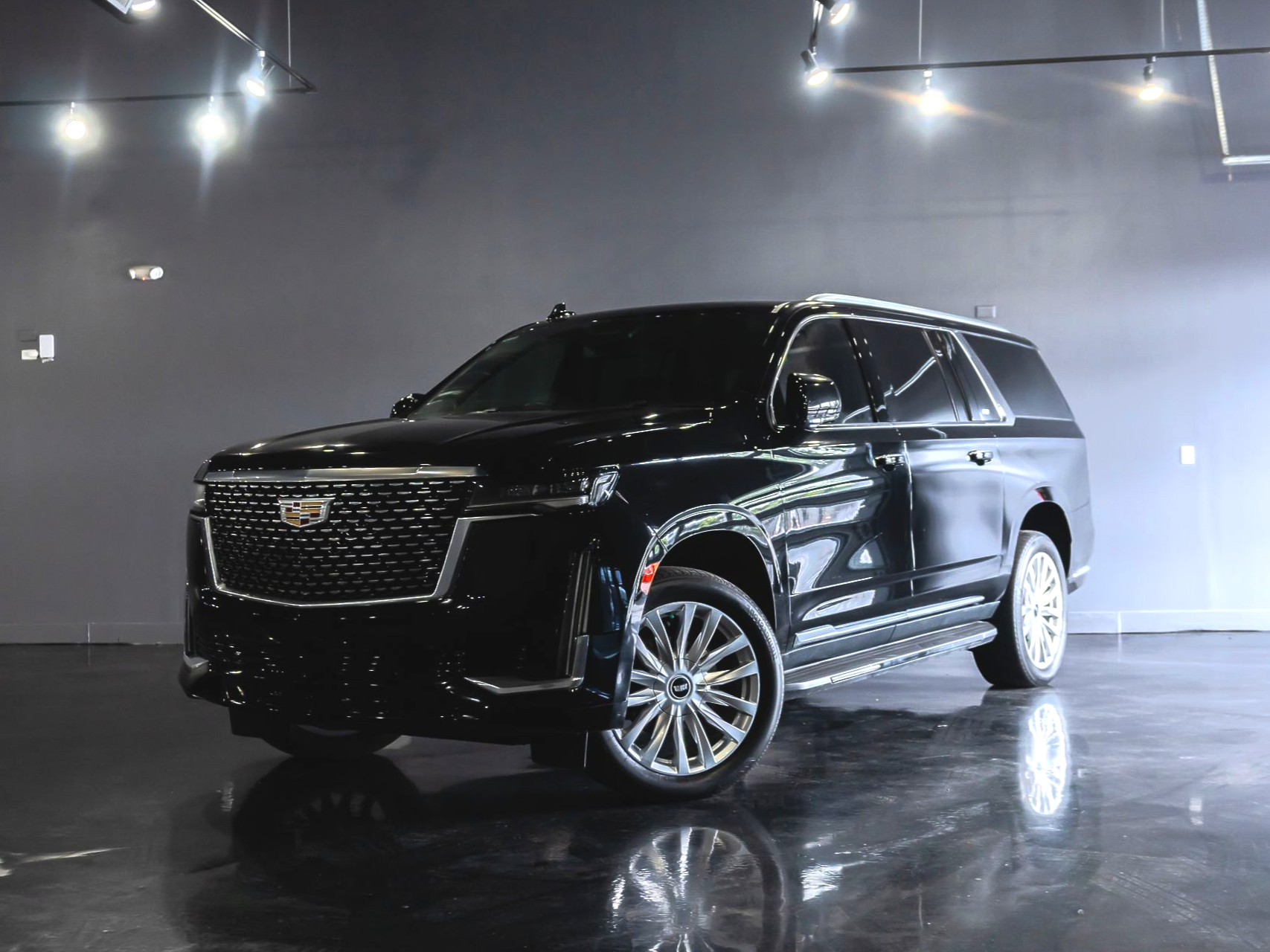 Black Cadillac Escalade SUV parked in a showroom, chrome wheels, reflective floor.