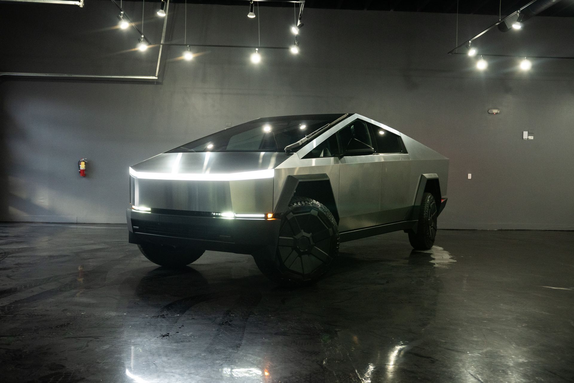 Tesla Cybertruck parked in a studio with lights overhead. Angular, silver vehicle.