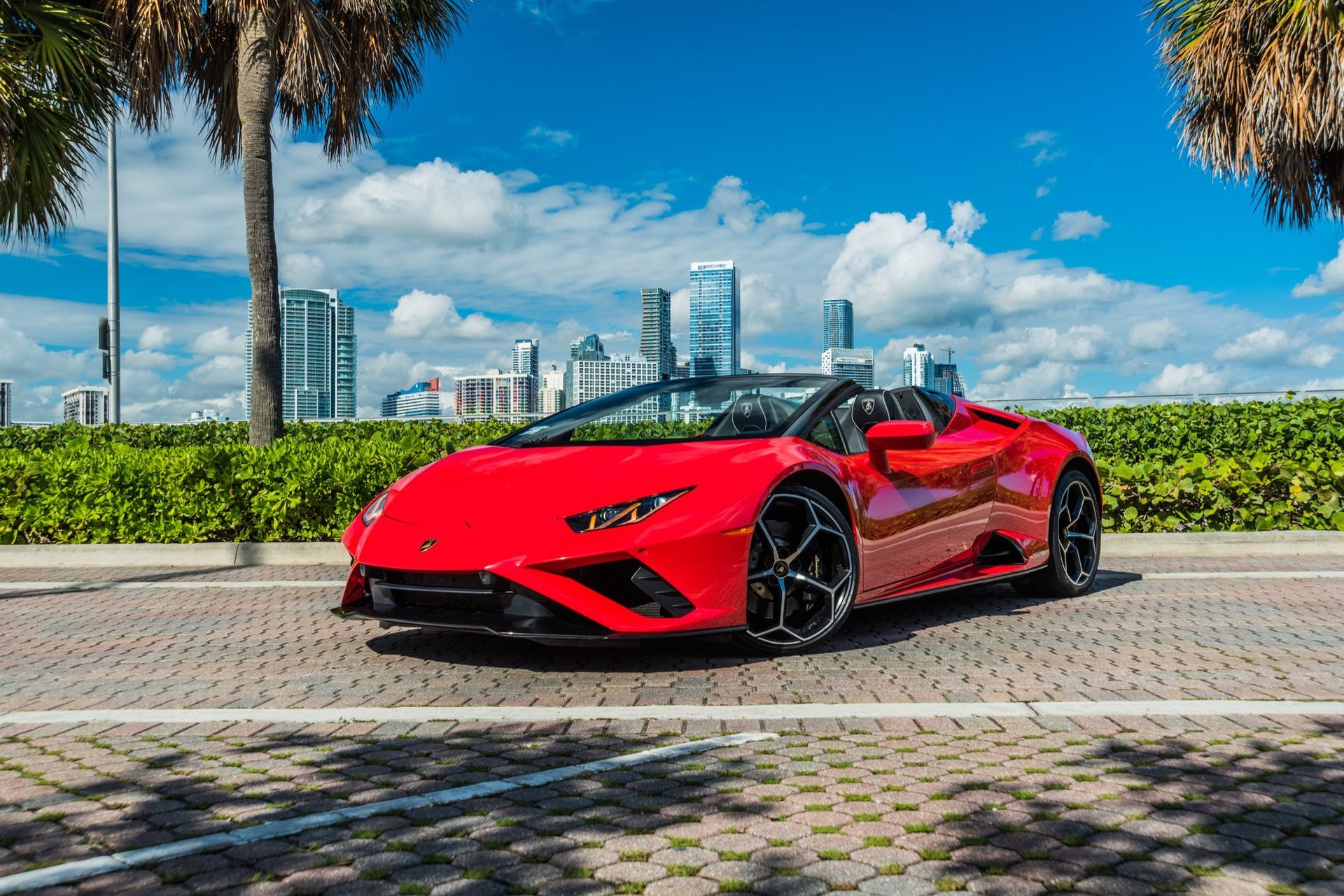 Red Lamborghini convertible parked on a paved street with a city skyline in the background.