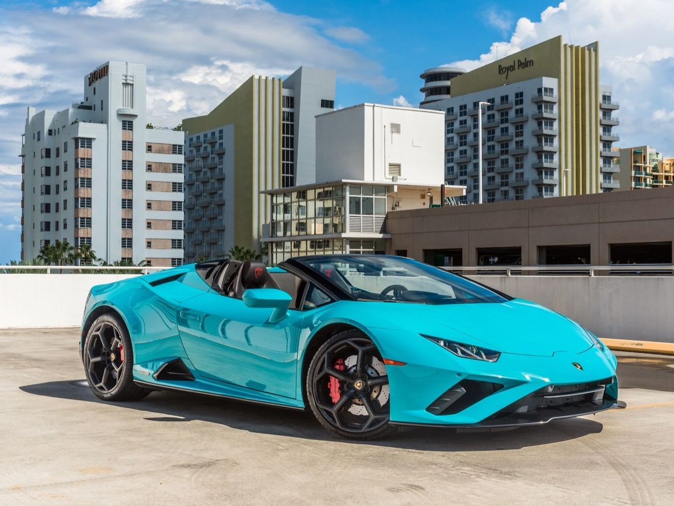 Teal Lamborghini convertible parked on a rooftop, with buildings and blue sky in the background.