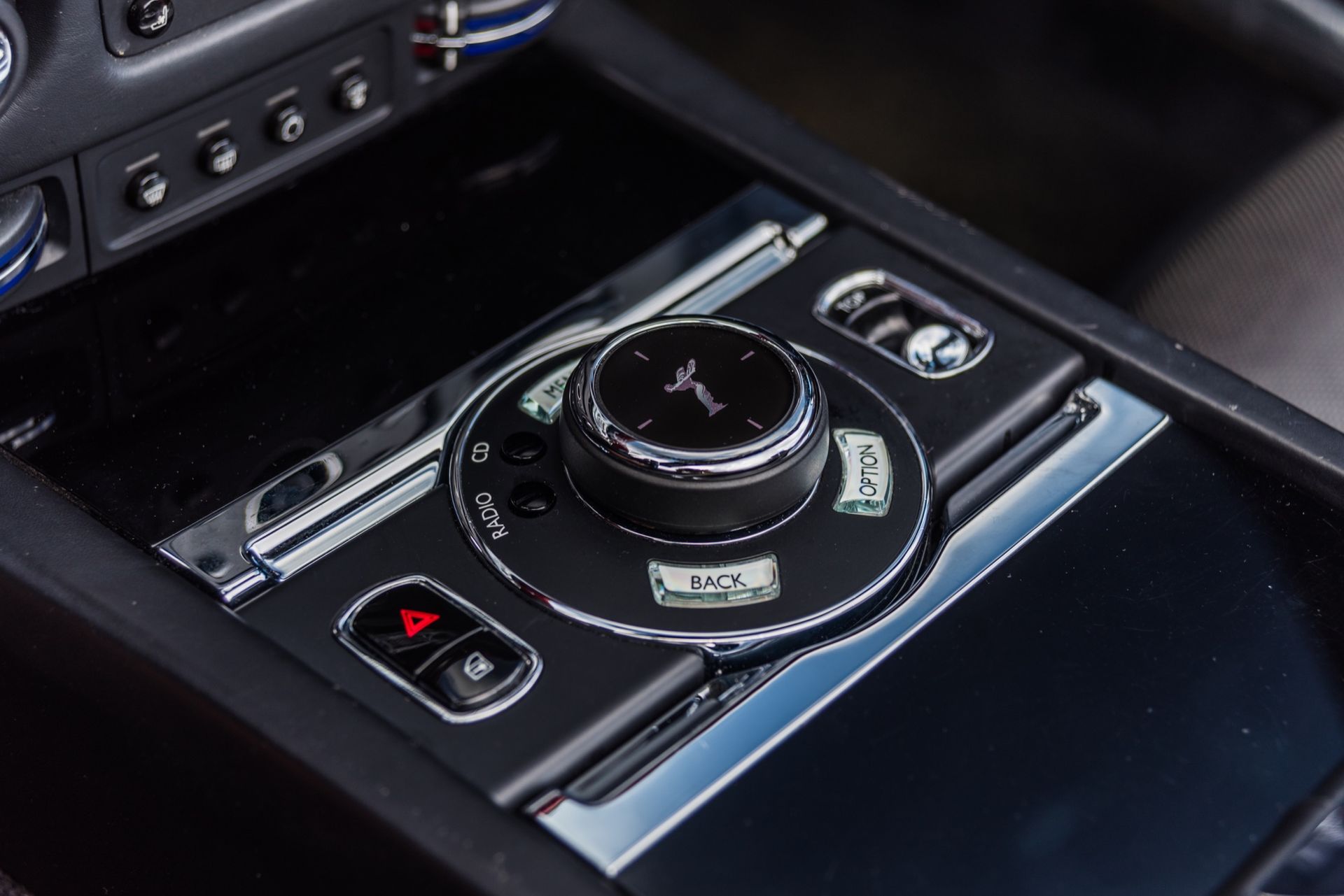Close-up of a car's control panel with a central dial for infotainment, chrome accents, and a warning button.