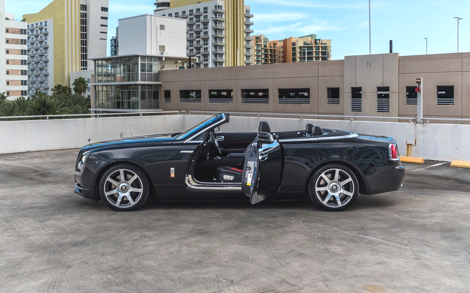 Dark Rolls-Royce convertible with open doors parked on a rooftop parking garage in front of city buildings.