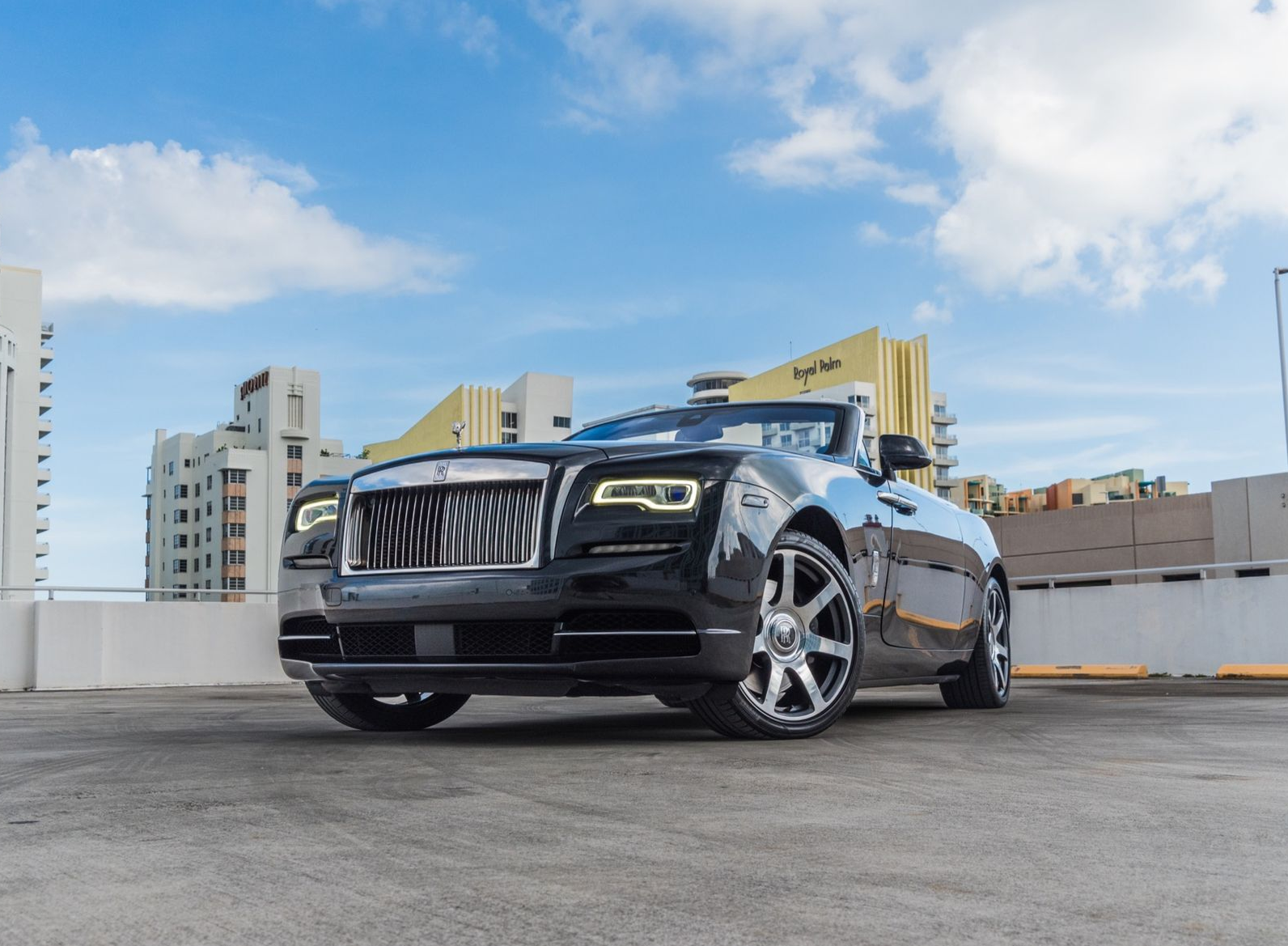 Black Rolls Royce convertible parked on a rooftop, against a blue sky with buildings.