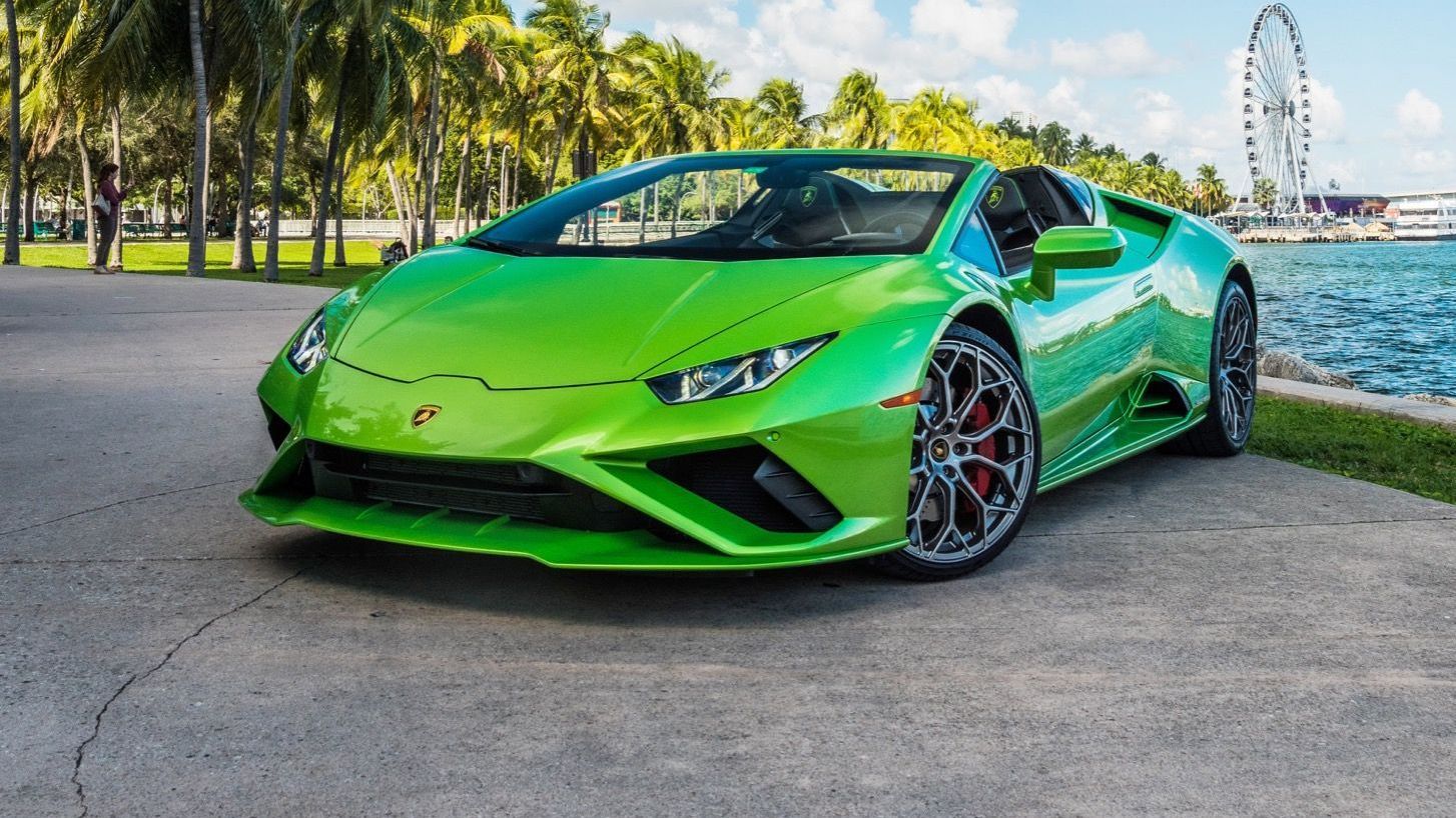 Green Lamborghini Huracan Spyder parked near water with a ferris wheel and palm trees in the background.
