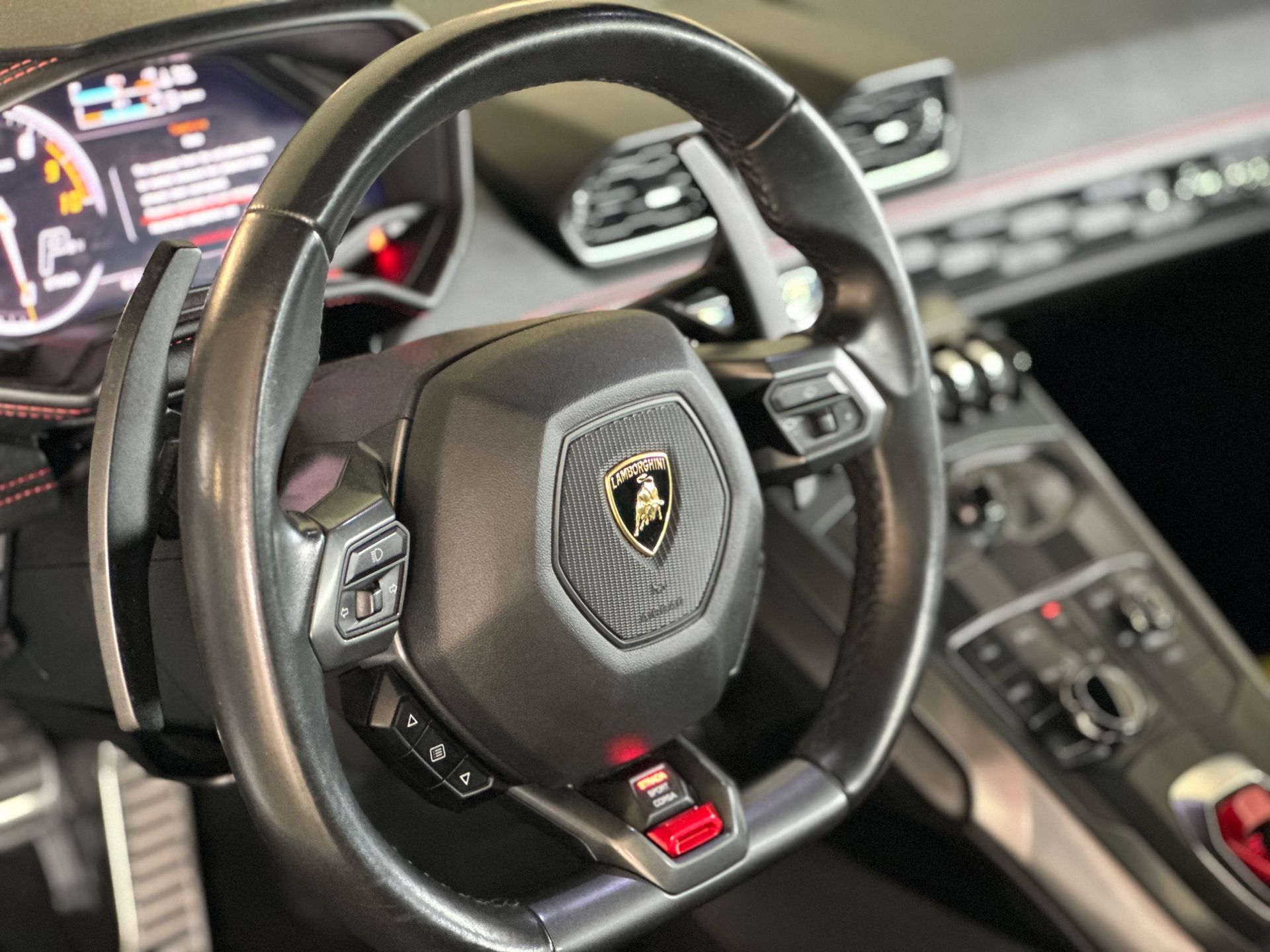 Lamborghini steering wheel, black leather, with controls and logo, in a car interior.