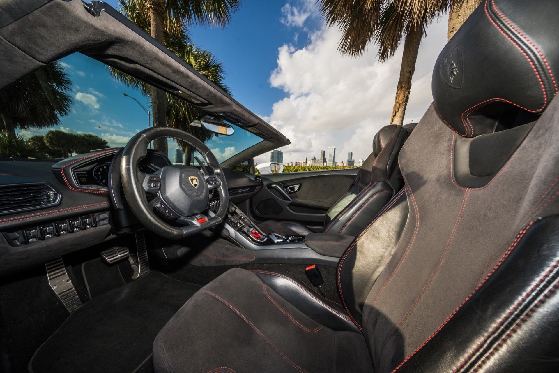 Interior of a black Lamborghini convertible with red stitching, parked under a blue sky.