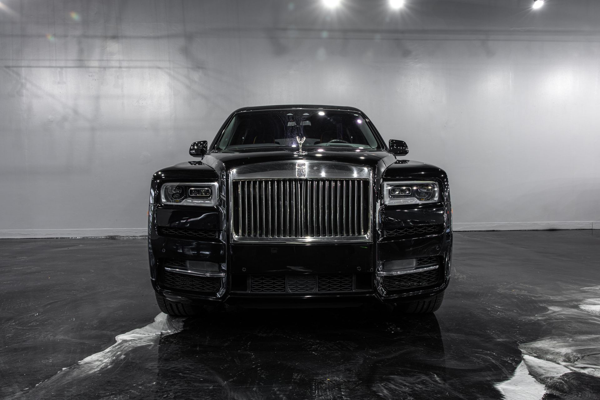 Black Rolls-Royce SUV, front view. Chrome grille, Spirit of Ecstasy hood ornament. Wet pavement, indoor setting.
