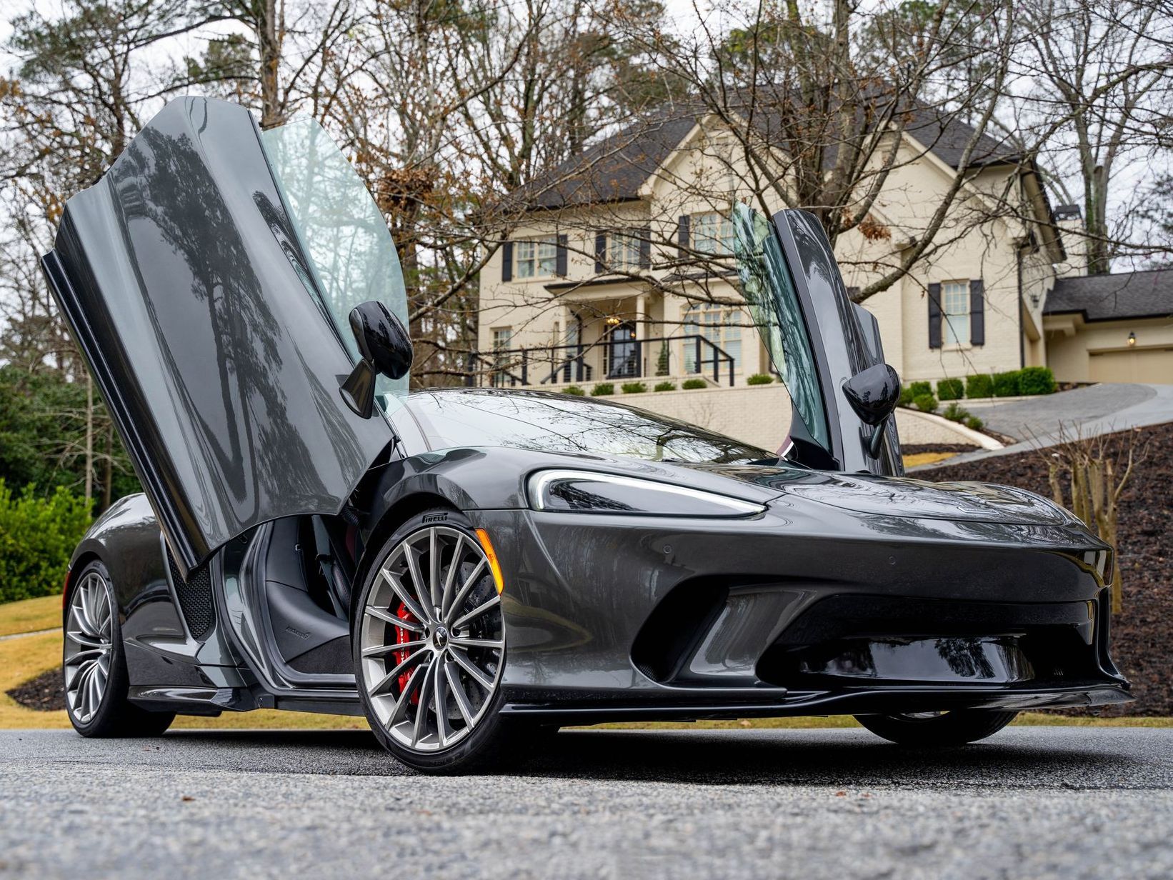 Gray McLaren sports car with doors open, parked on a driveway, with a house in the background.