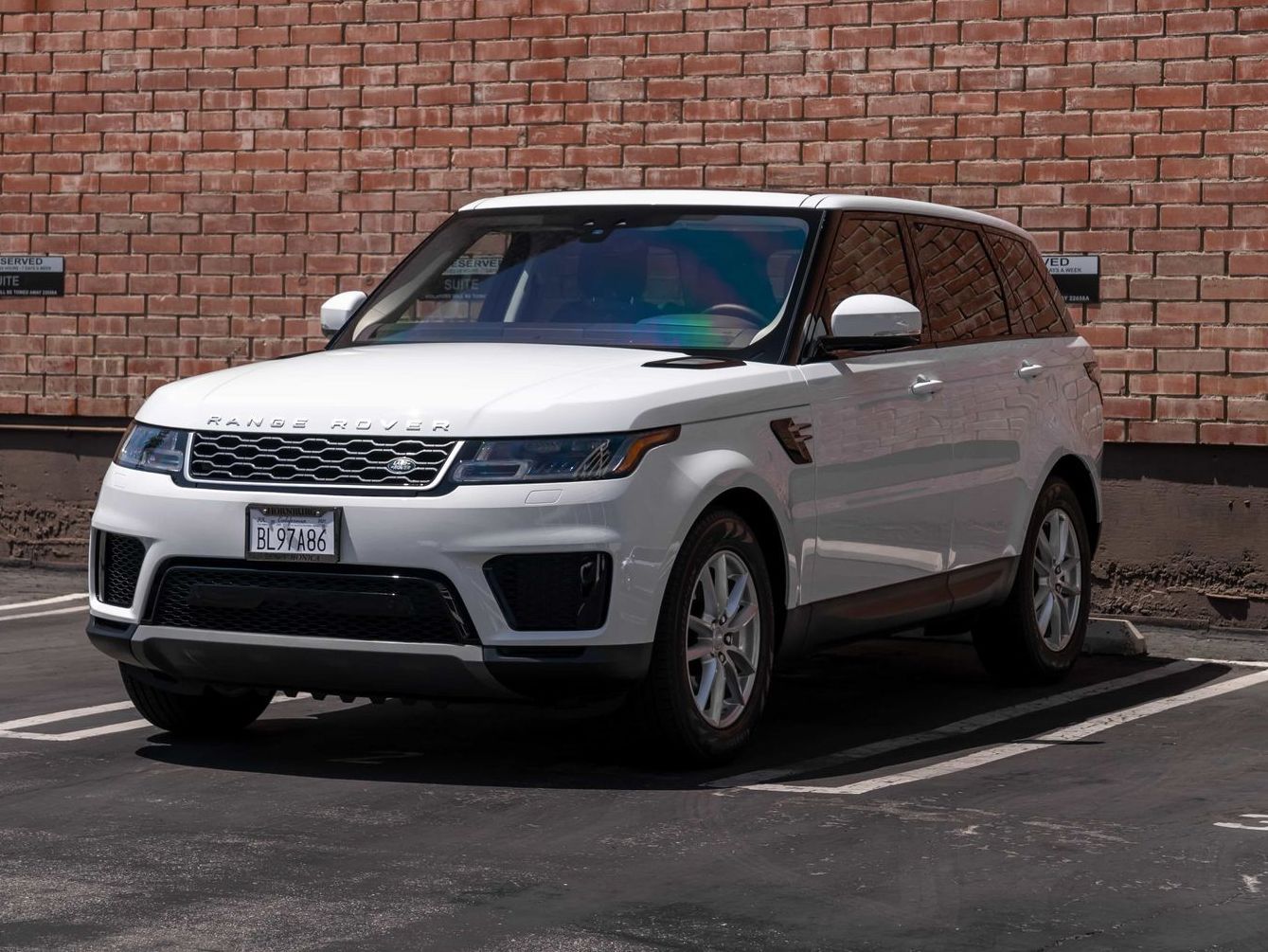 White Range Rover Sport SUV parked in front of a brick wall in a parking lot.
