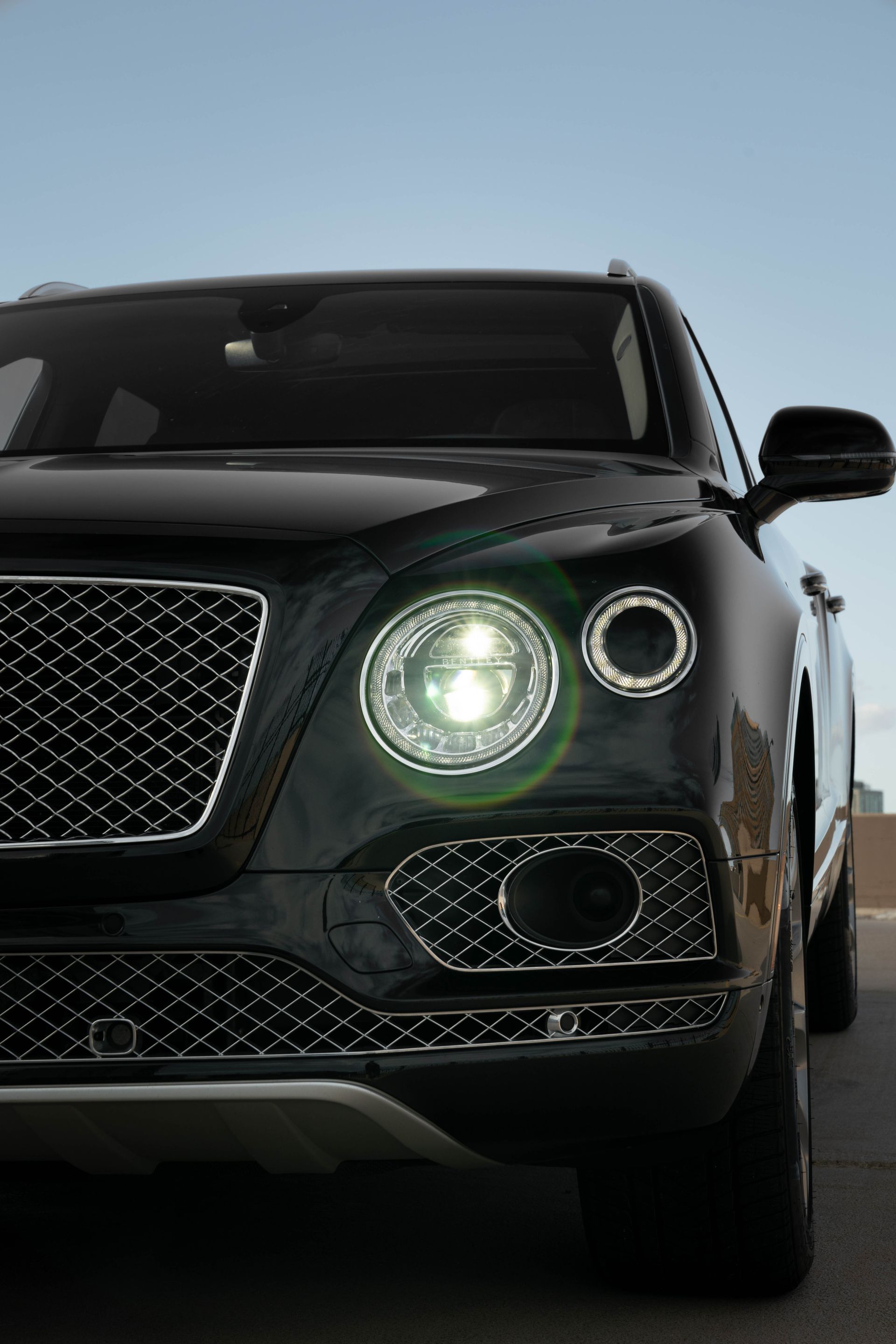 Black Bentley SUV, front view. Bright headlights, chrome grille, parked outside.