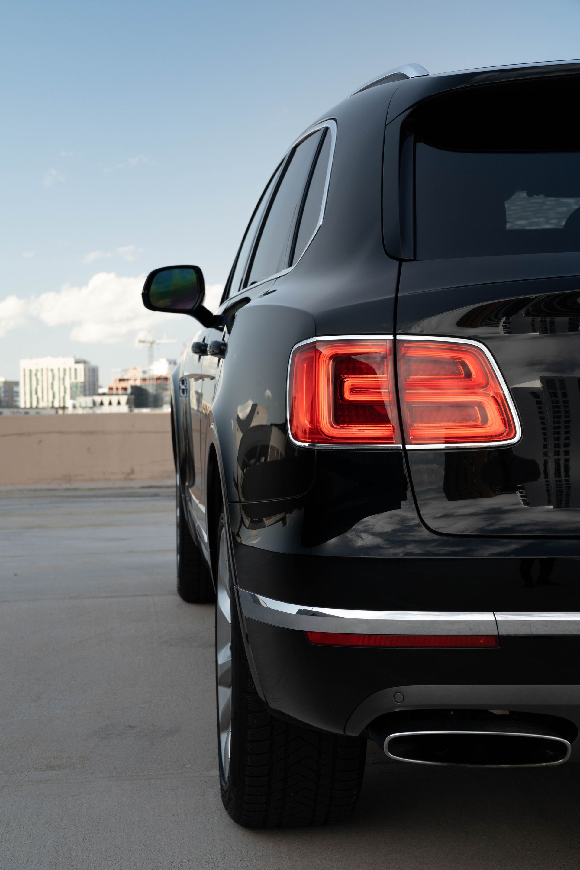 Black Bentley SUV, rear view, on a concrete surface, with buildings and sky in background.