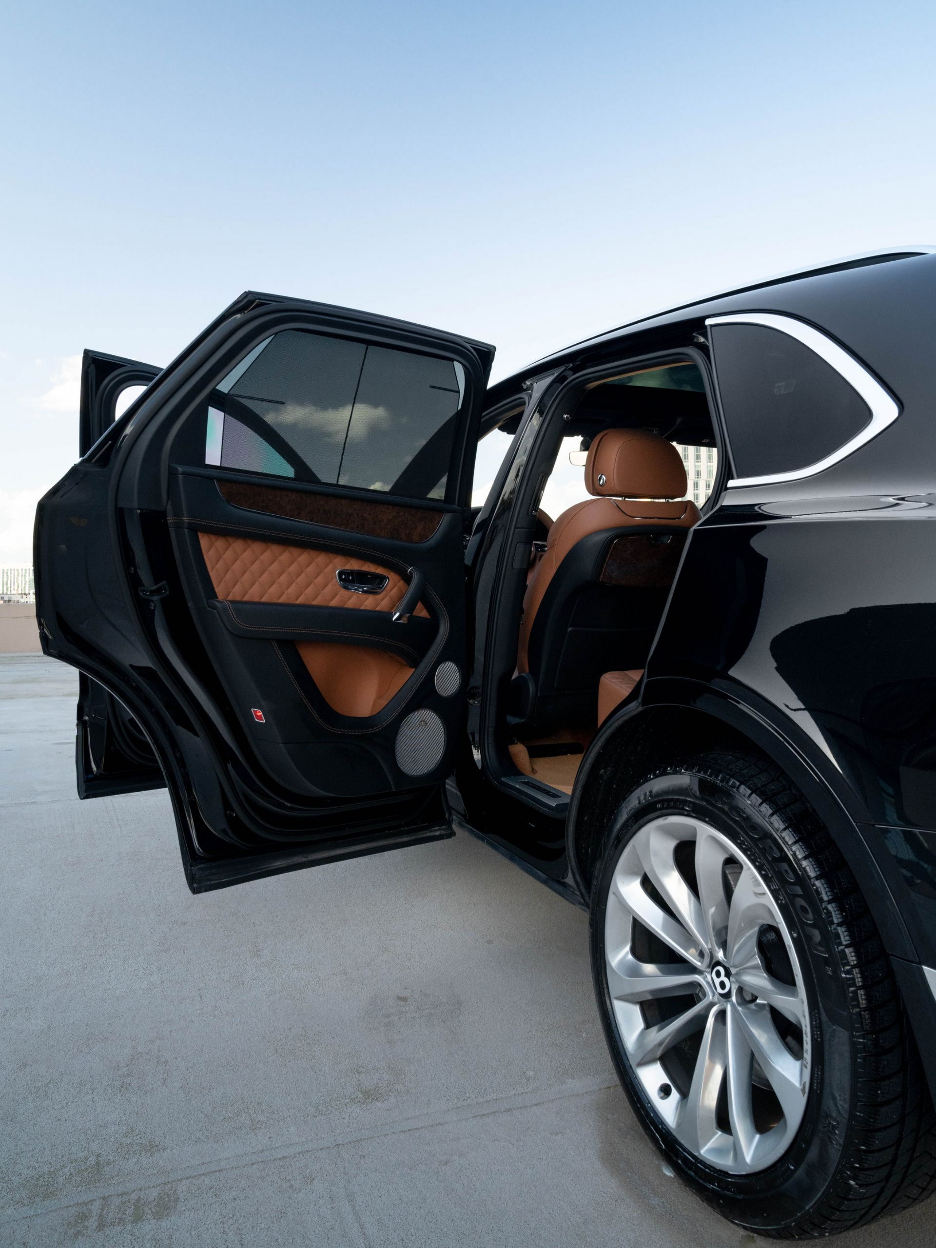 Black luxury SUV with open doors, tan leather interior, silver wheels, against a light blue sky.