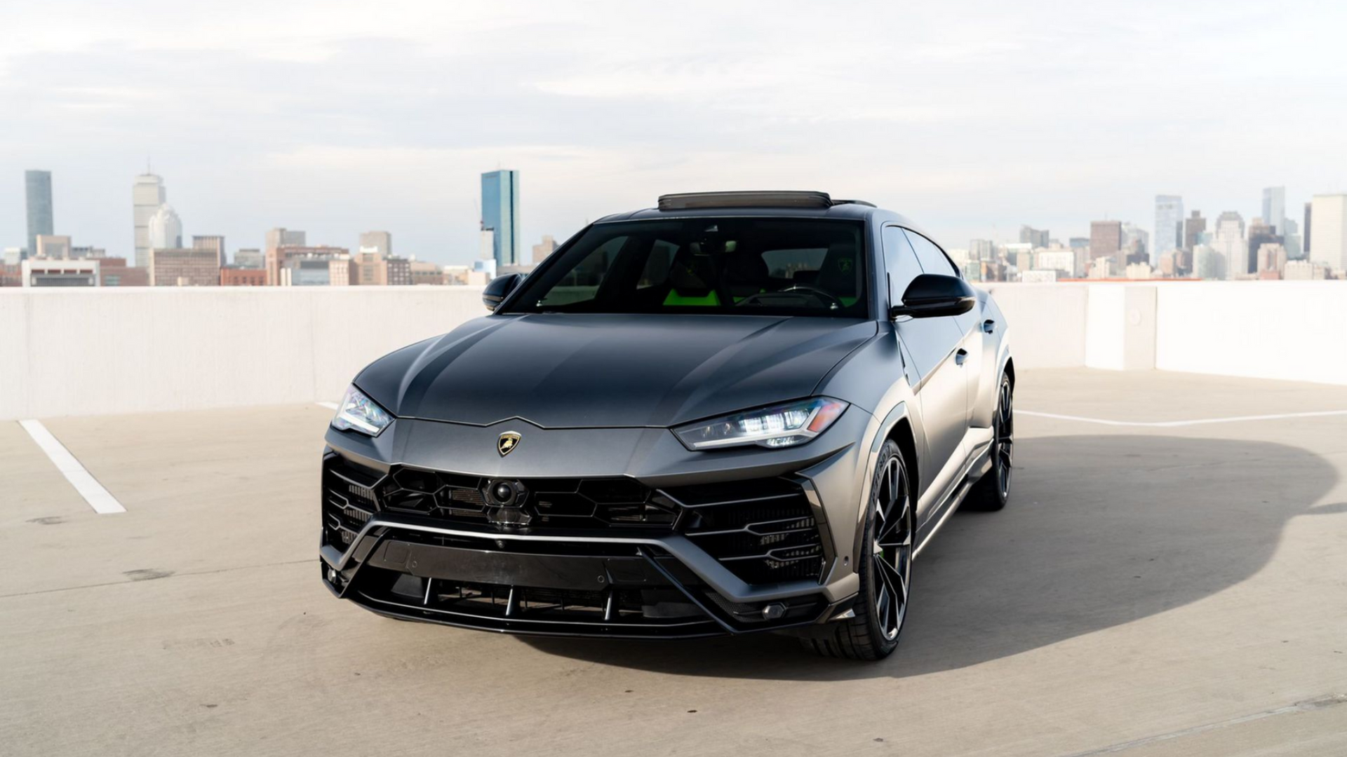 Dark gray Lamborghini Urus SUV parked on a rooftop, cityscape in the background.