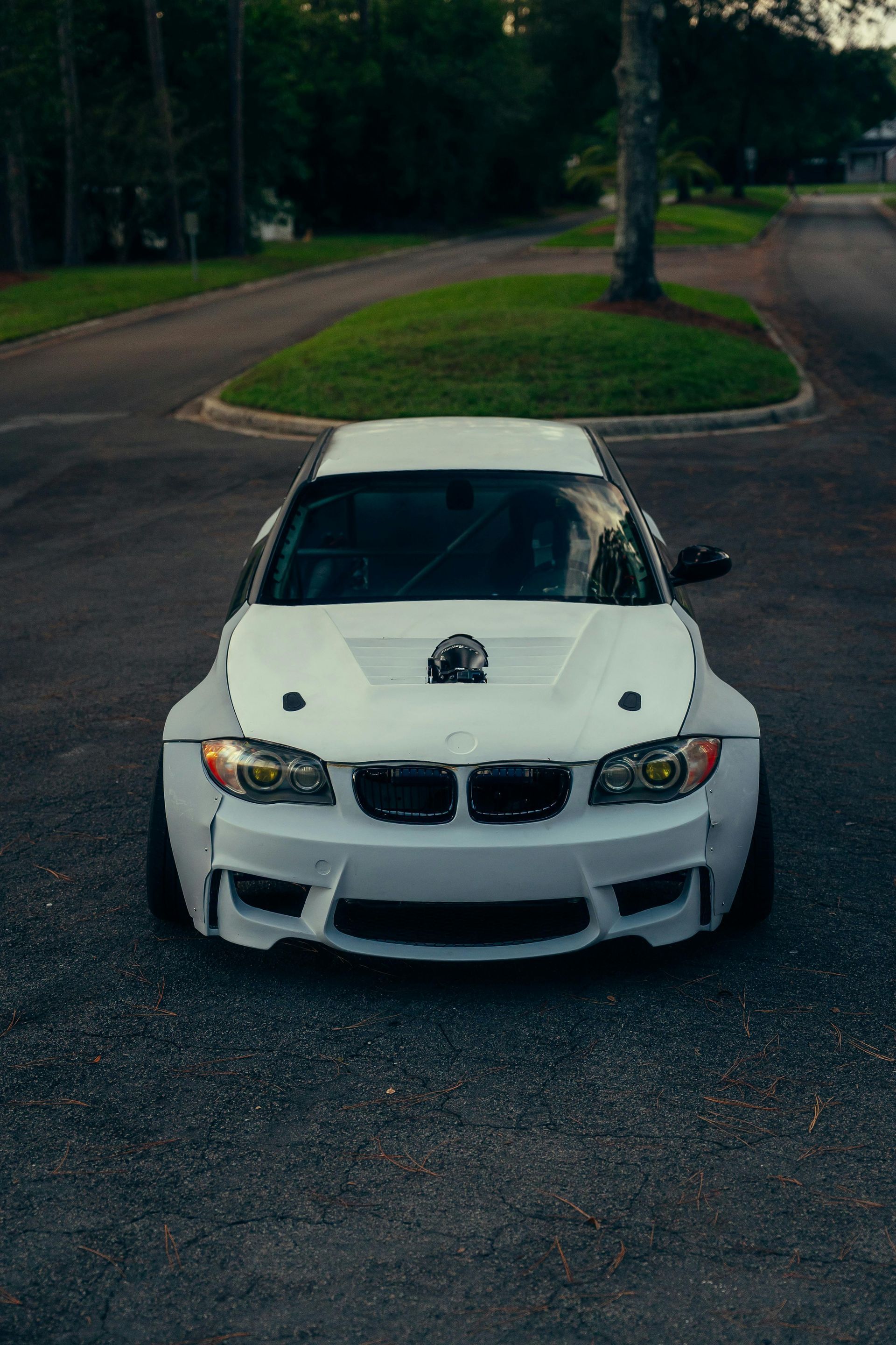 White modified BMW car on asphalt, hood scoop visible, wide body kit, in a driveway.