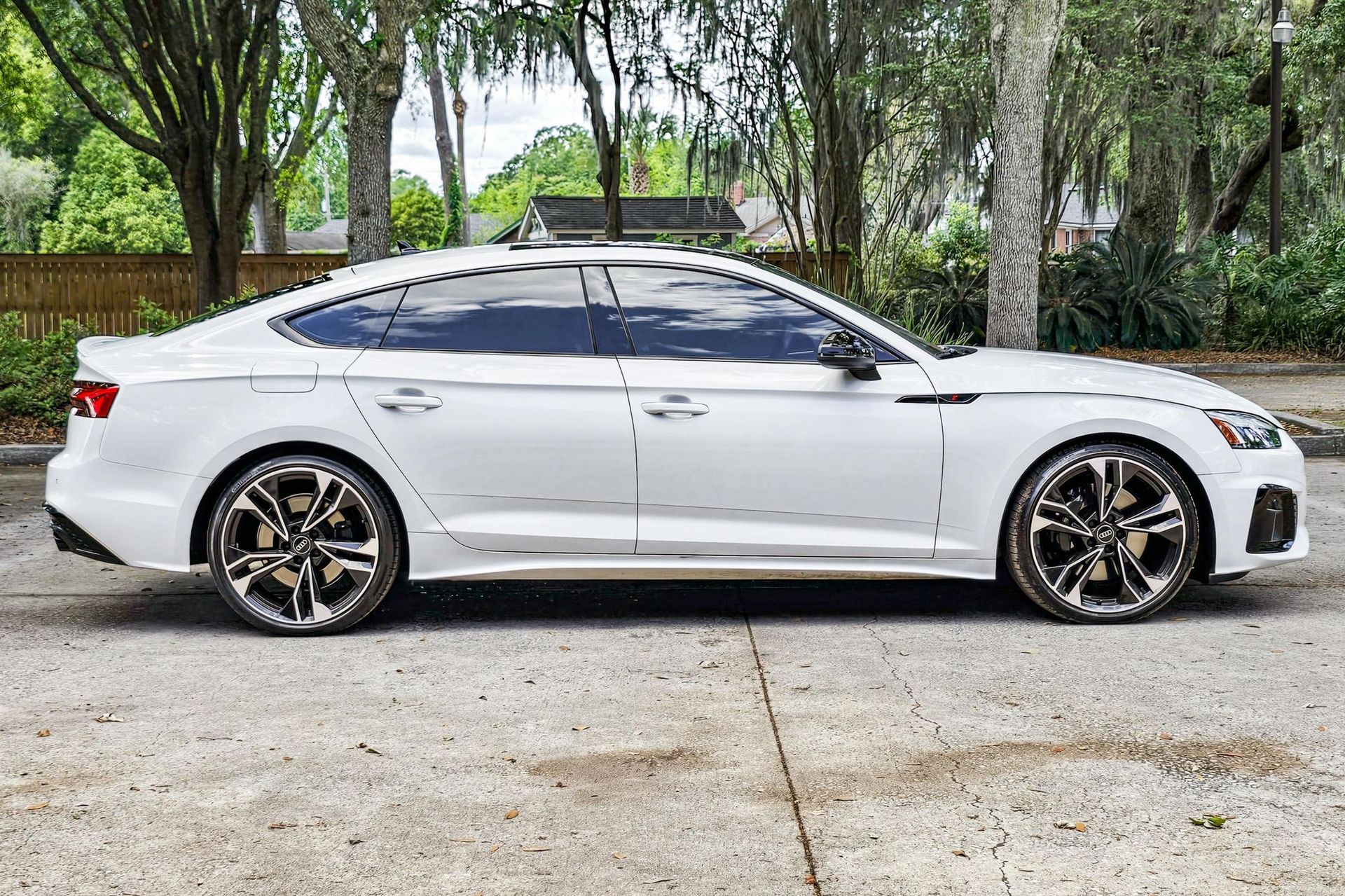 White Audi sedan parked on a paved road; tinted windows, dark rims, trees in background.