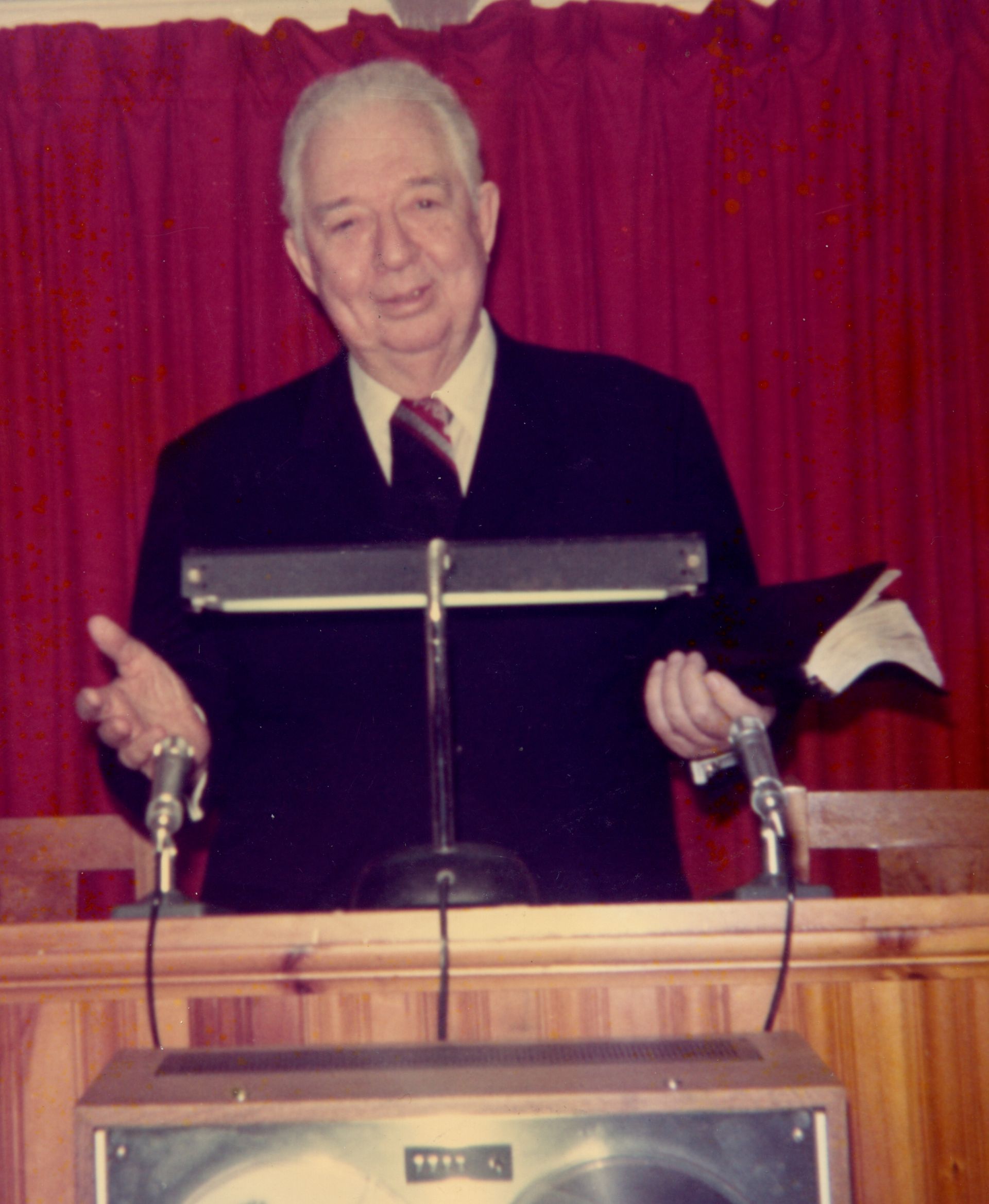An older man in a suit speaking behind a wooden podium with two microphones and a red curtain backdrop.