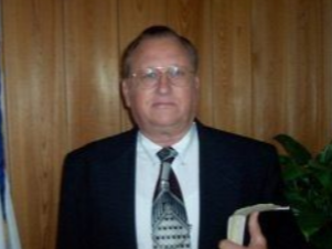 A person wearing a suit and patterned tie, holding a Bible in front of a dark-paneled wall.