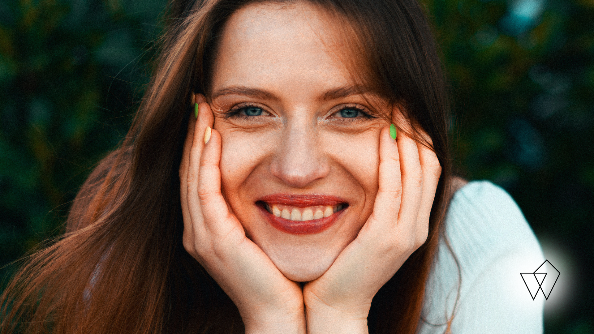 Woman smiling, hands on cheeks, long brown hair, green painted nails, natural setting.