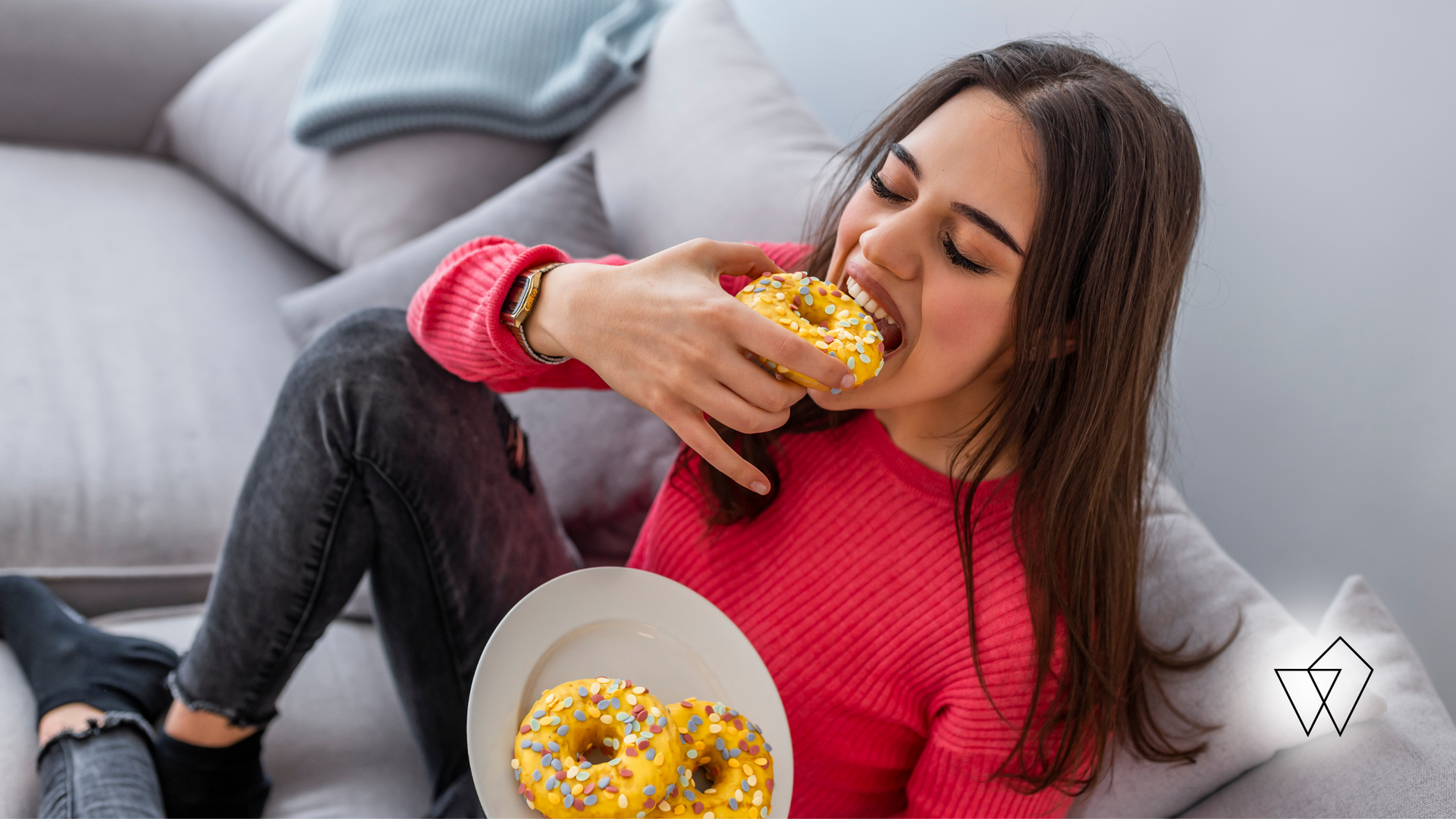 Woman on couch eating a donut, holding a plate of donuts. She wears a pink shirt, black jeans.