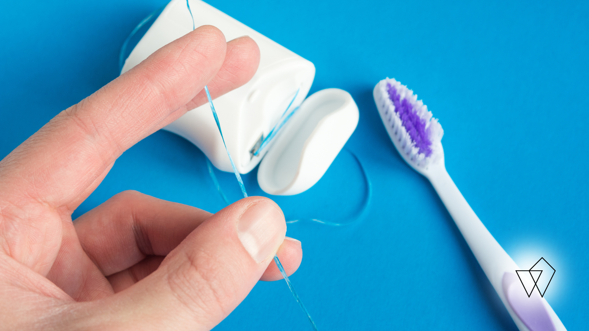 Hand holding a dental floss threader next to a toothbrush and floss container on a blue background