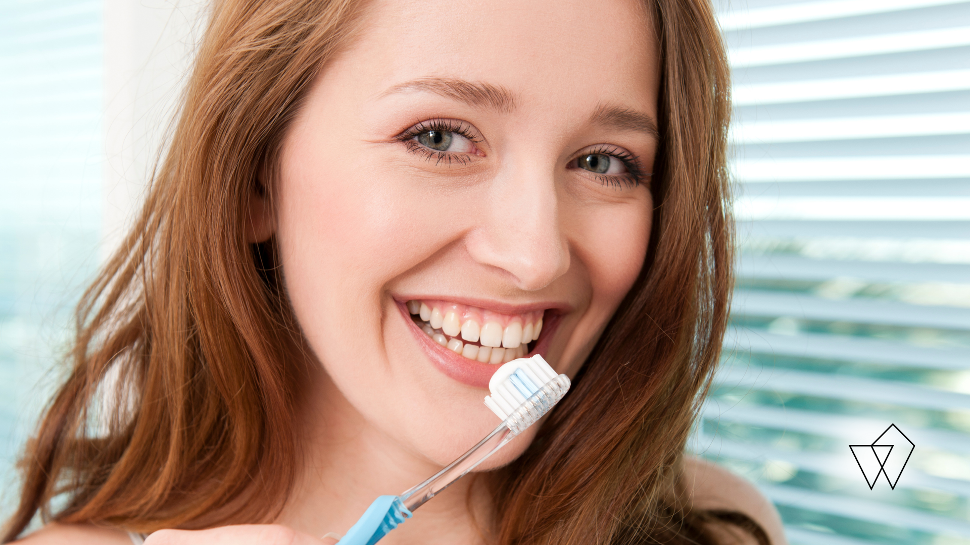 Woman smiling, holding a toothbrush with toothpaste, ready to brush her teeth, near a window.