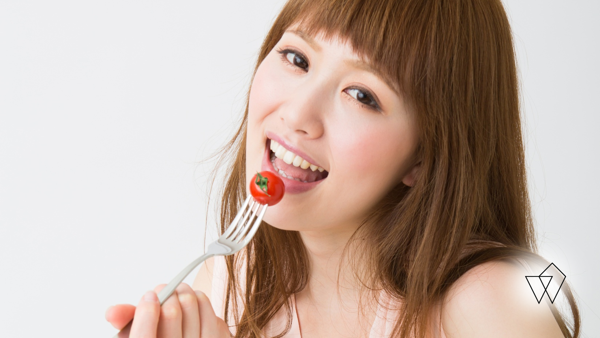 Woman with brown hair eats a cherry tomato from a fork, smiling.