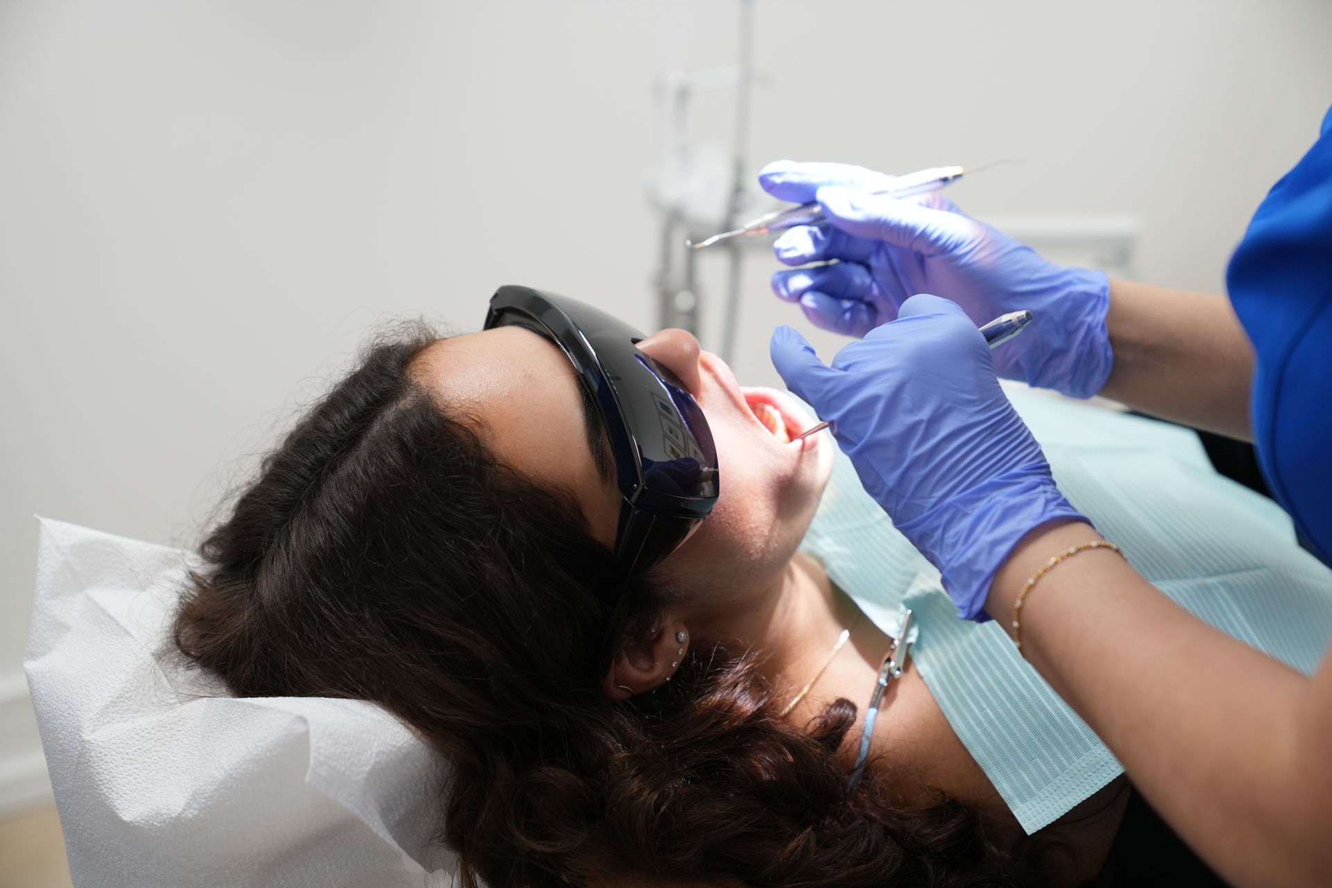 Teen with braces at a dentist, mouth open, being examined with tools. Gloves, white coat, and blue chair.