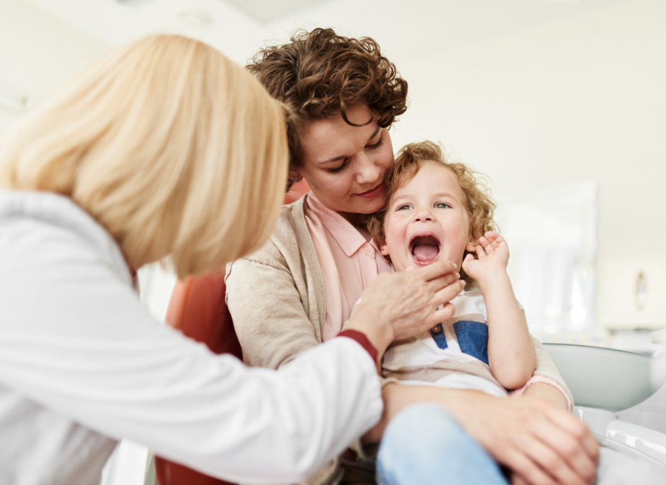 Dentist examining a patient's teeth. Assistant holding tools. All wearing masks, gloves, and scrubs.