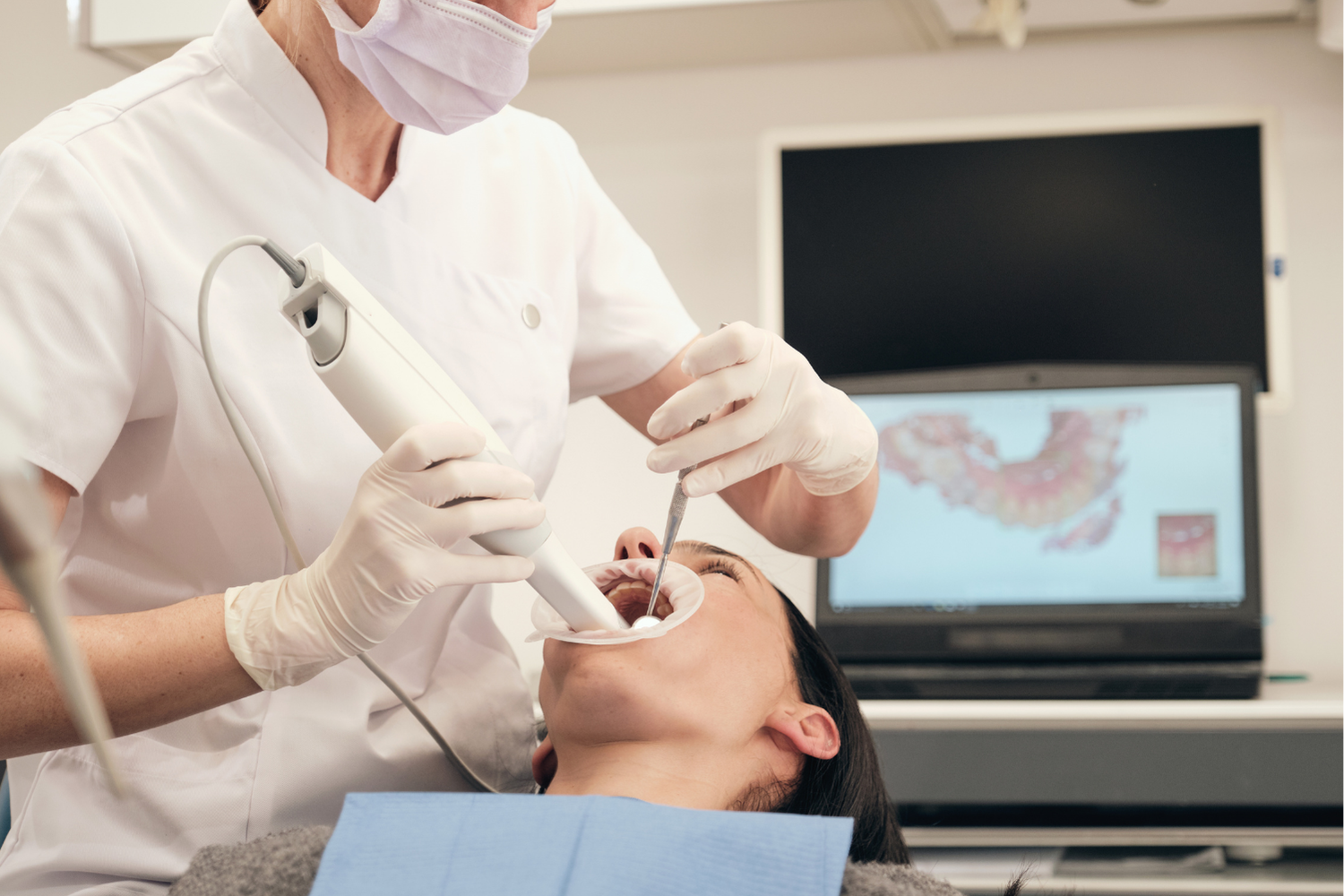 Dentist scans a patient's open mouth with an intraoral scanner, screen shows dental scan in a clinic.