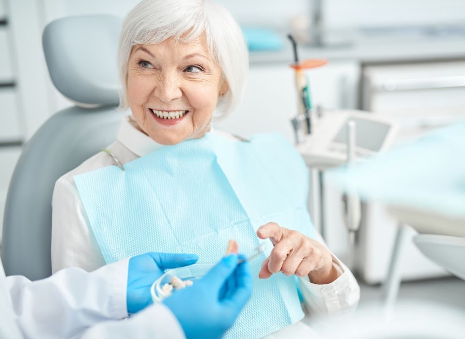 A woman smiles, sitting in a dental chair, looking at a dental appliance held by someone wearing gloves.