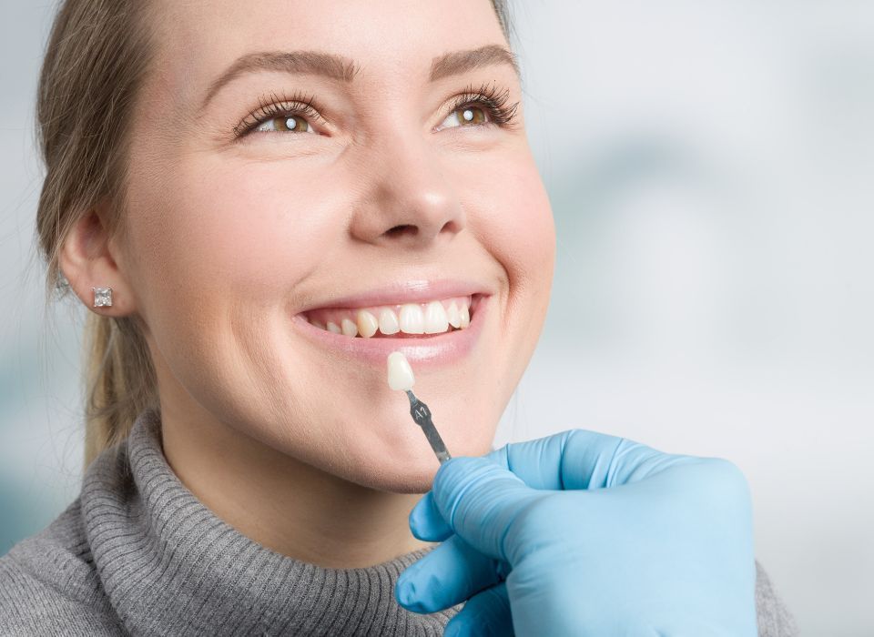 Woman smiling, comparing a tooth shade guide with teeth, held by a person wearing a blue glove.