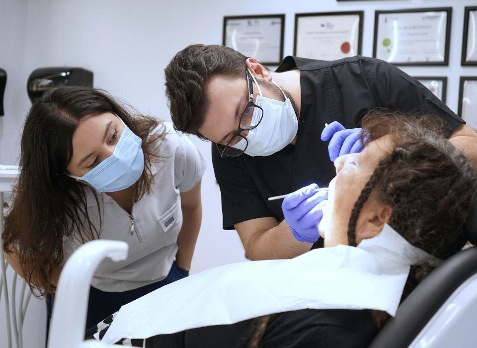 Dentist and assistant examining patient's teeth in a dental office. Both wear masks and gloves.