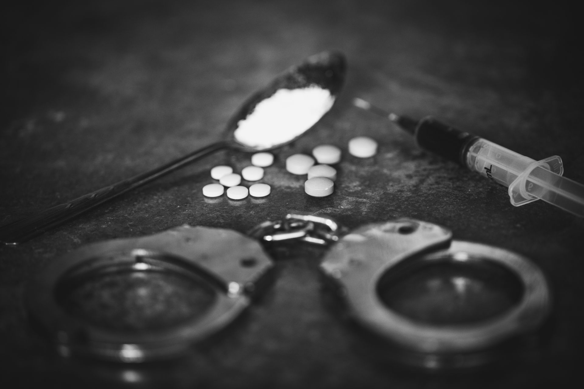 A black and white photo of handcuffs , a syringe , a spoon , and pills.