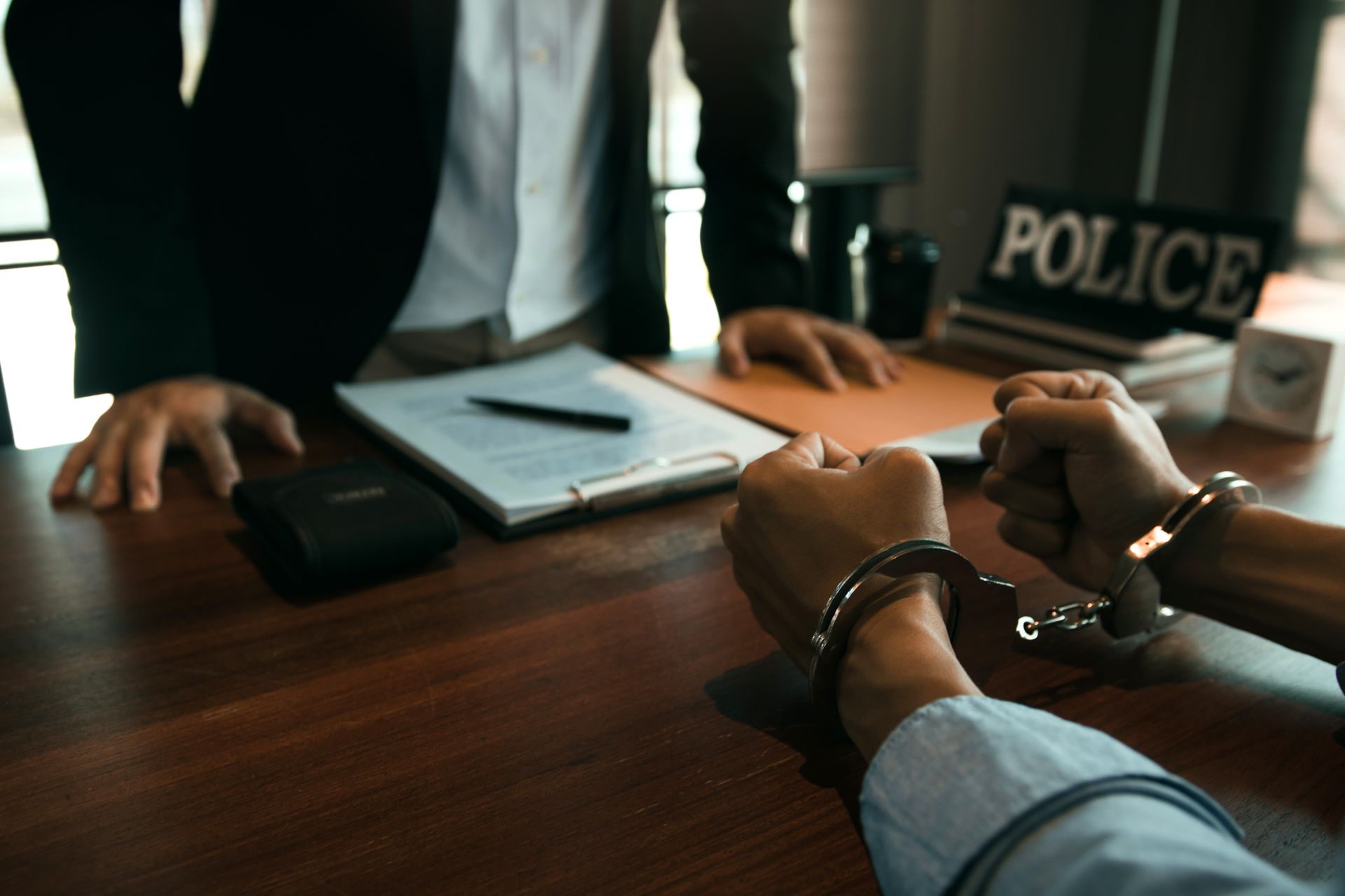 Person in handcuffs at a desk facing a person in a suit, a police sign in the background.