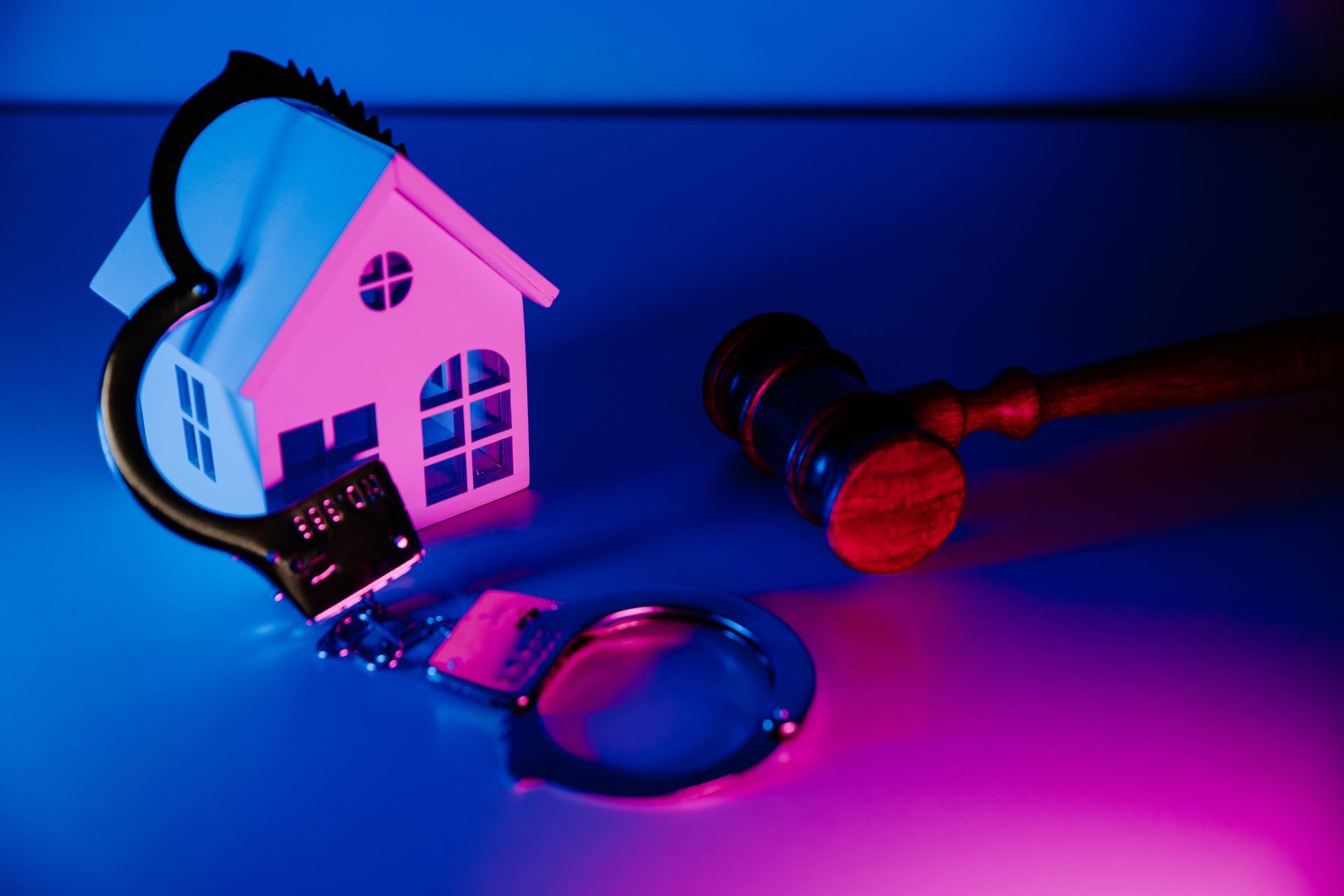 A paper house with handcuffs, next to a gavel, under blue and pink light.