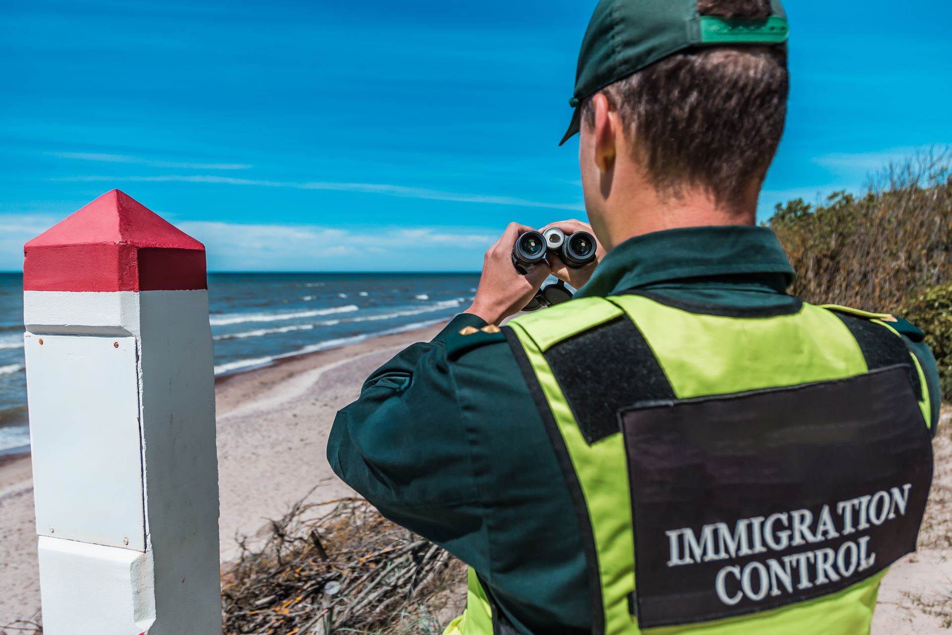 Border patrol officer at the coast, looking through binoculars; white & red marker, blue sky, ocean.